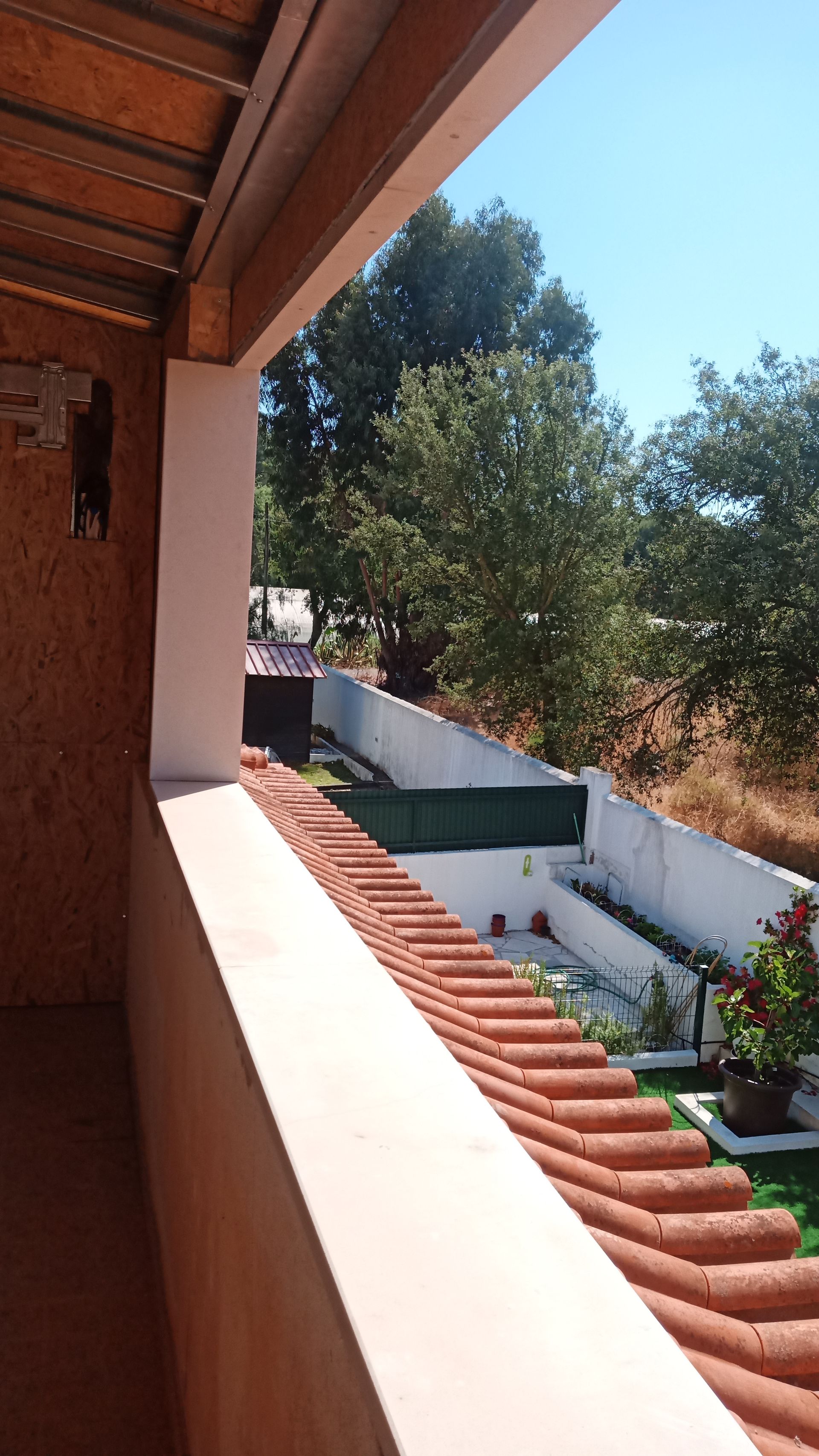 Vista de uma varanda coberta com telhado de telhas vermelhas, com vista para um jardim arborizado e céu azul.