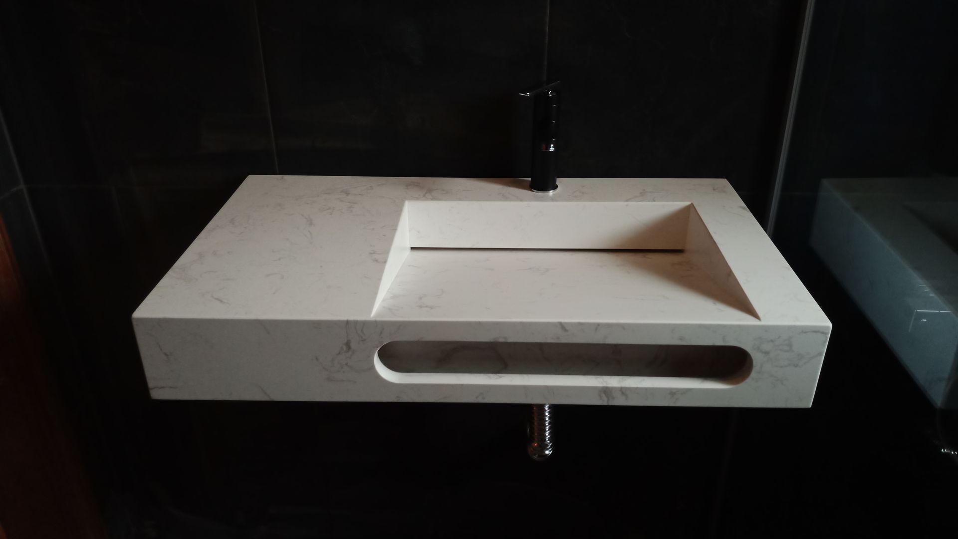 Pia retangular de pedra bege, fixada na parede, com torneira preta, em uma parede revestida com azulejos pretos.