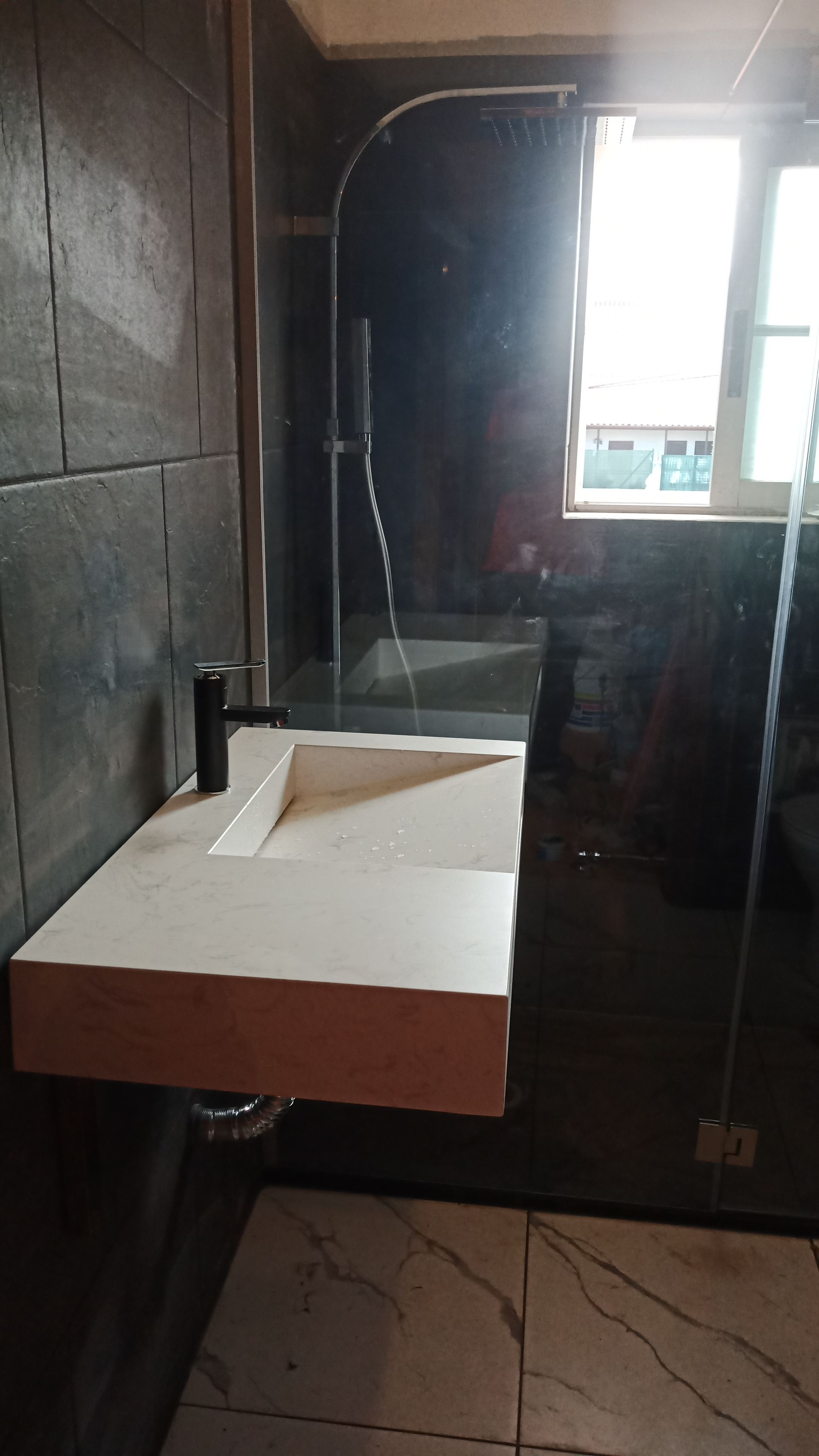 Banheiro com pia suspensa branca, torneira preta e box de vidro para o chuveiro.