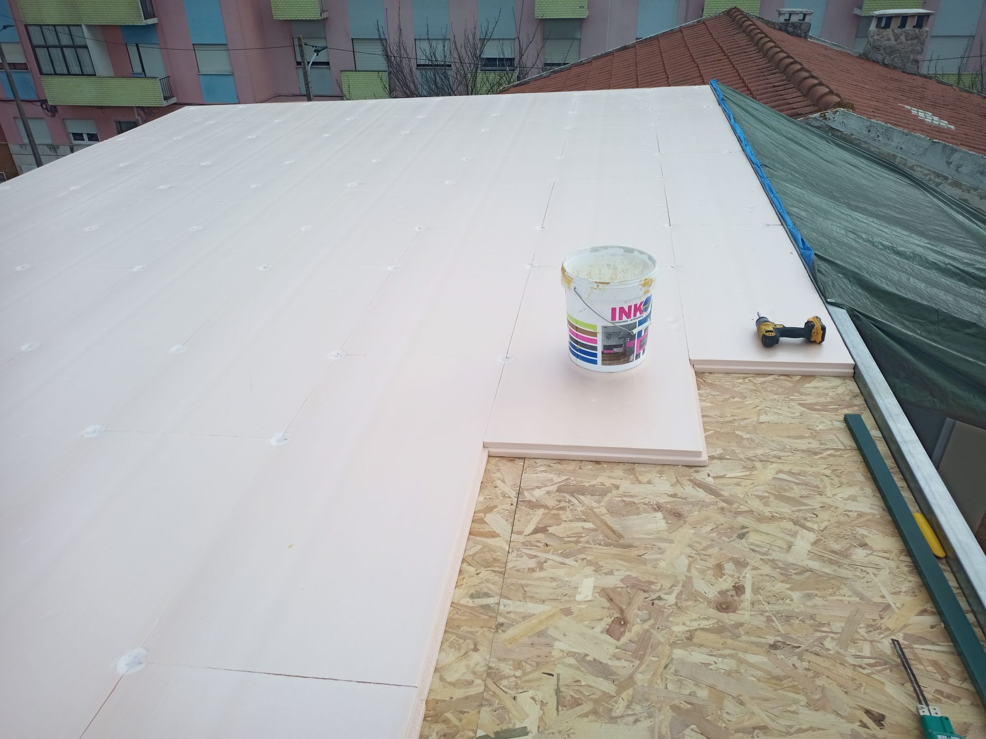 Instalação de isolamento de telhado com painéis de espuma rosa sobre revestimento OSB; um balde e ferramentas estão próximos.
