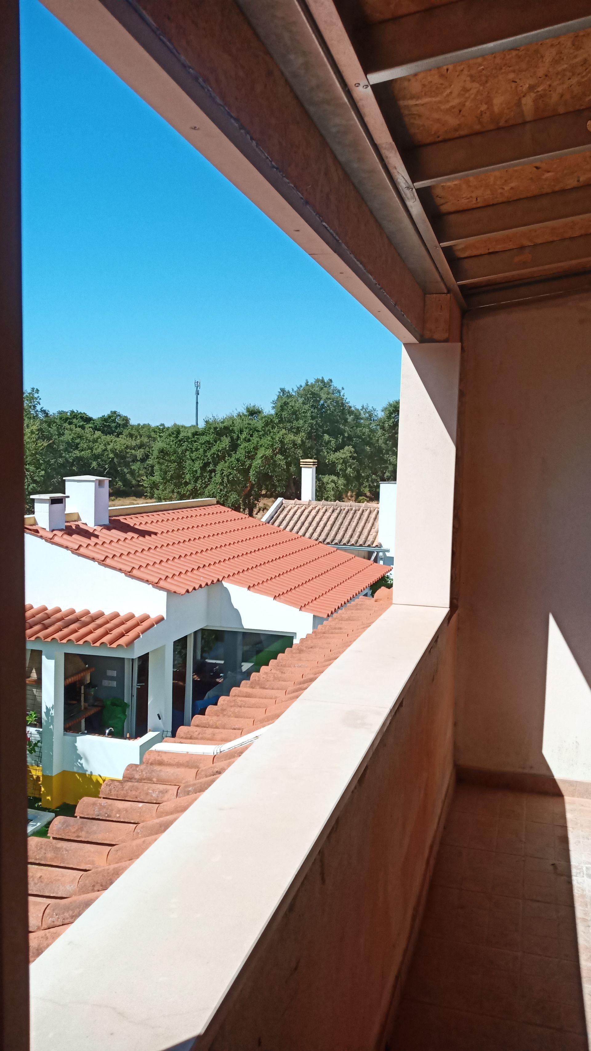 Vista de telhados de telha vermelha, árvores e céu azul a partir de uma varanda.