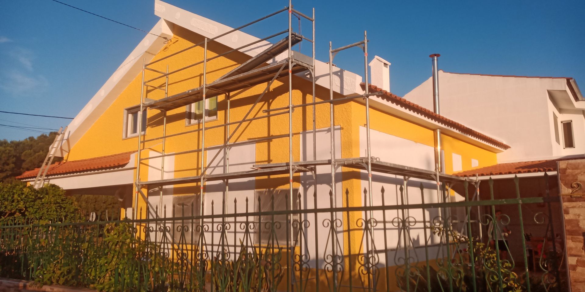 Casa amarela com andaimes montados para reforma. Uma cerca de metal e folhagem estão em primeiro plano.