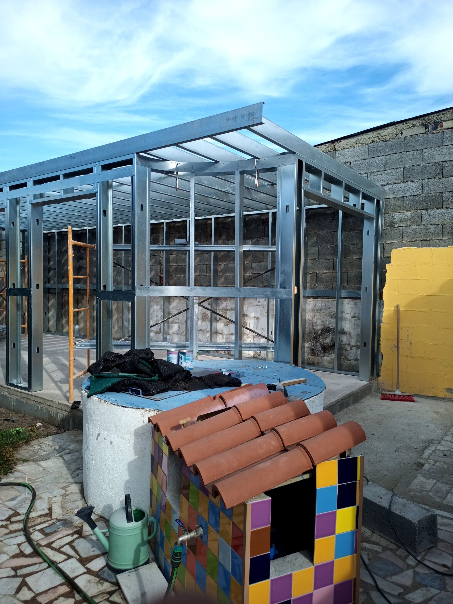 Estrutura metálica de uma varanda envidraçada em construção perto de um muro de pedra, com uma pequena casinha de brincar colorida na frente.