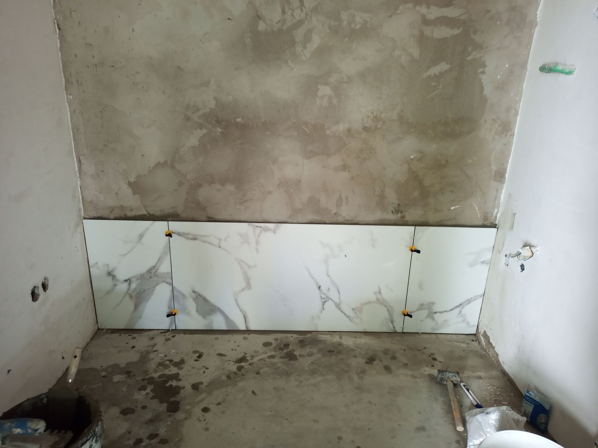 Azulejos sendo instalados em uma parede durante a construção; azulejos com estampa de mármore cinza e branco são visíveis.