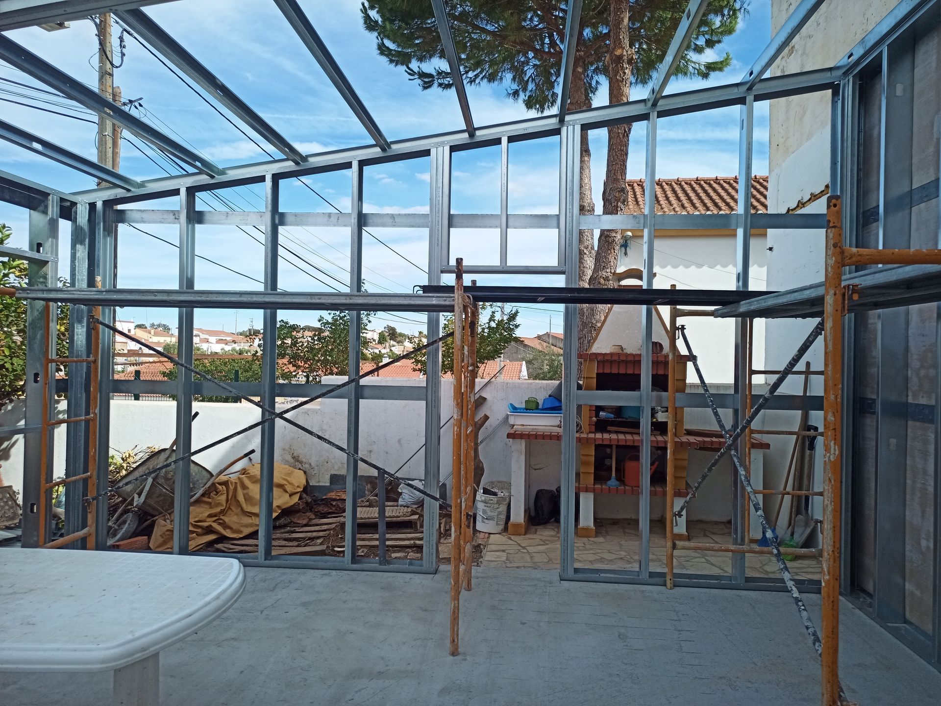 Estrutura metálica de uma varanda envidraçada com teto de vidro, situada em um pátio com vista parcial para uma casa.