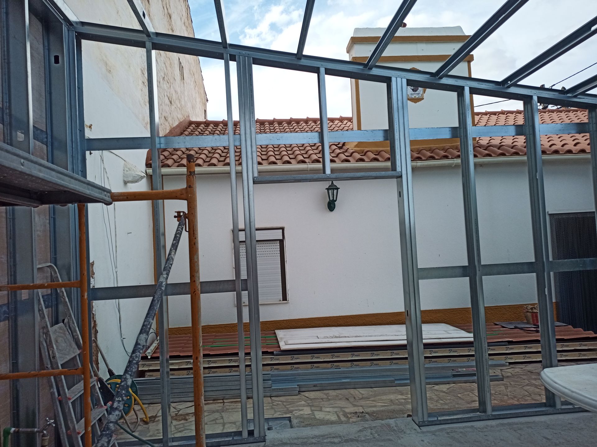 Estrutura metálica de uma construção em andamento; ambiente externo com paredes brancas e telhado de telhas vermelhas visíveis ao fundo.