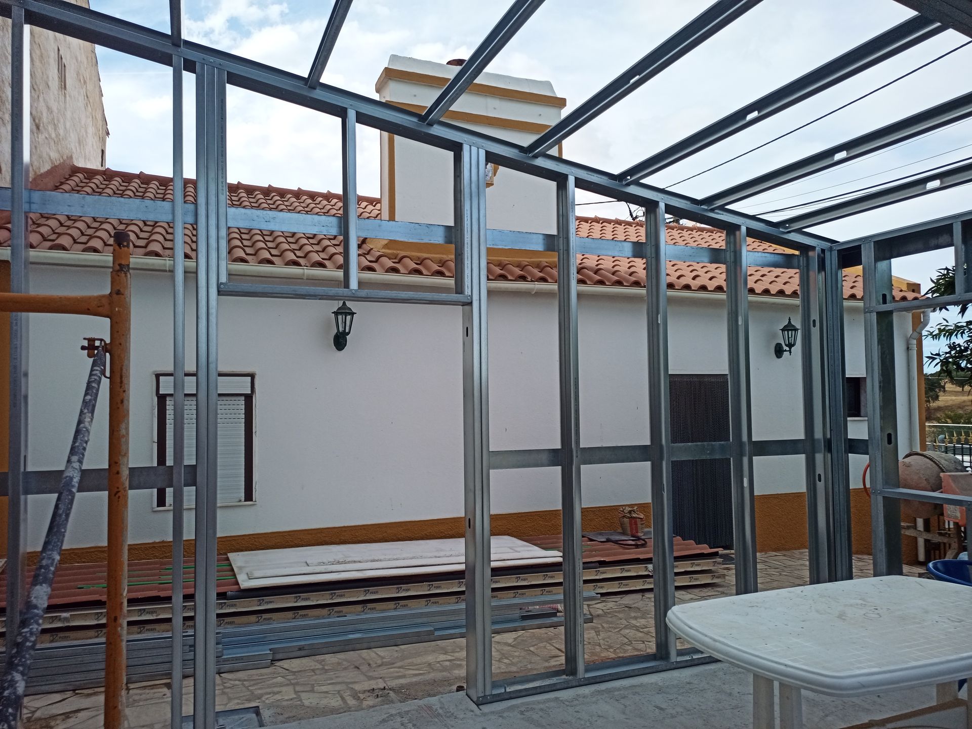 Estrutura metálica de uma construção externa, com um edifício branco e telhado de telha vermelha ao fundo.