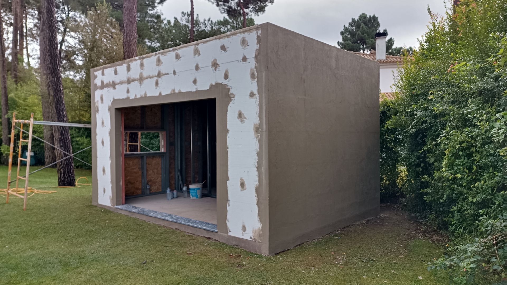 Pequeno prédio em construção, exterior em estuque, porta aberta, rodeado por grama verde e árvores.