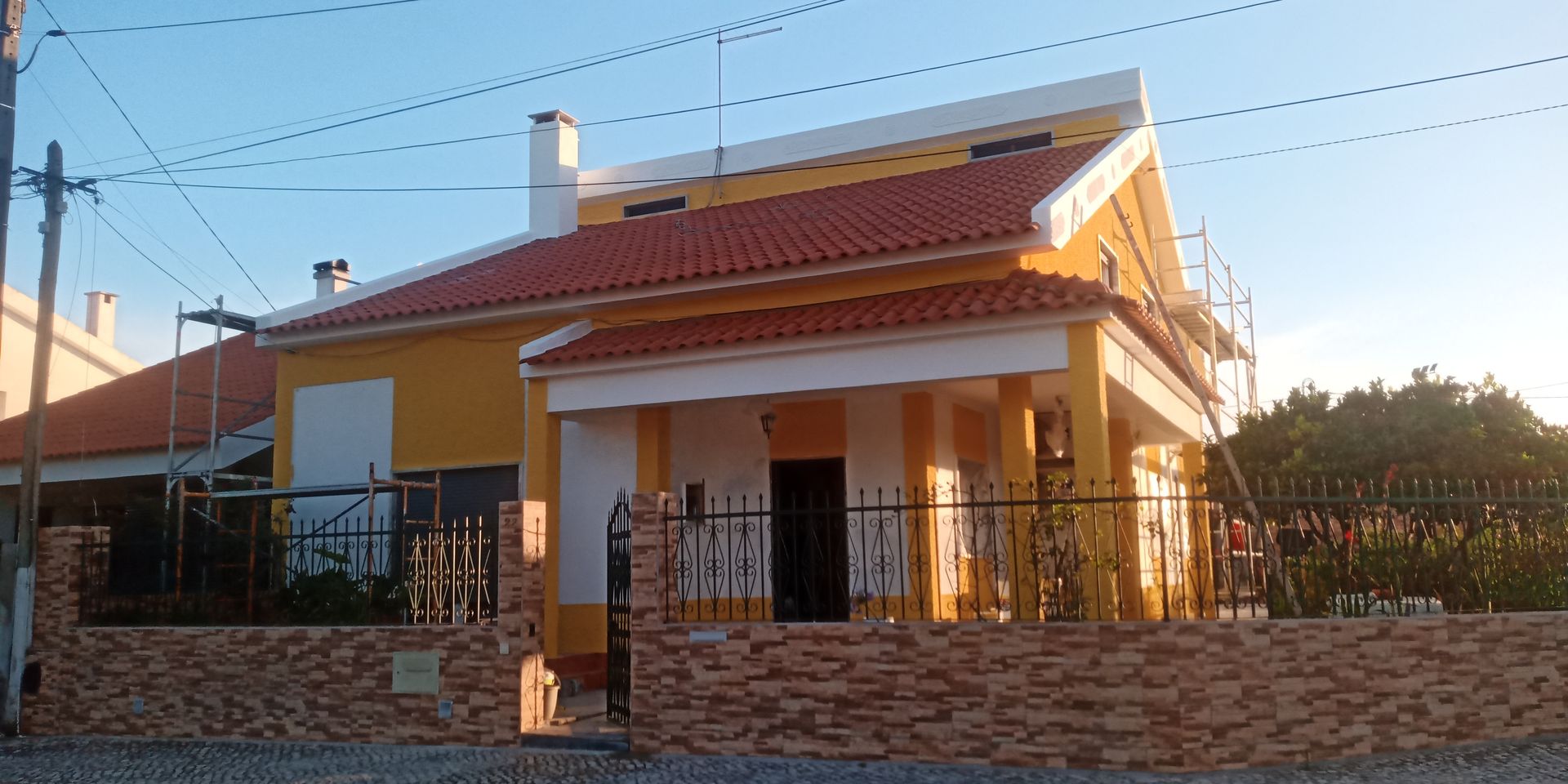 Casa amarela com telhado de telha vermelha e detalhes brancos, atrás de um muro de tijolos e um portão preto.
