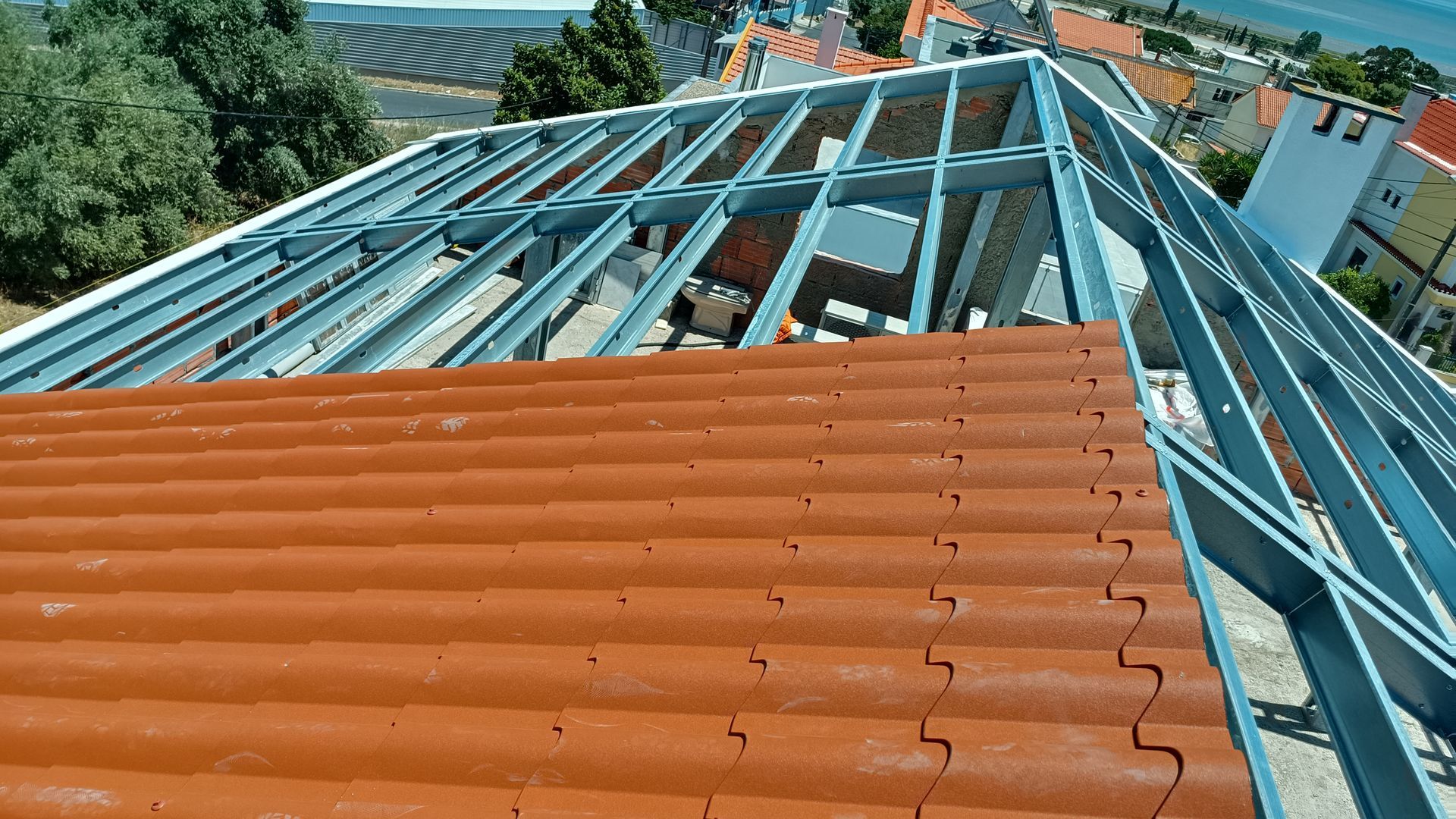 Um telhado em construção, mostrando telhas vermelhas sendo colocadas sobre uma estrutura metálica, com vista para prédios e árvores ao fundo.