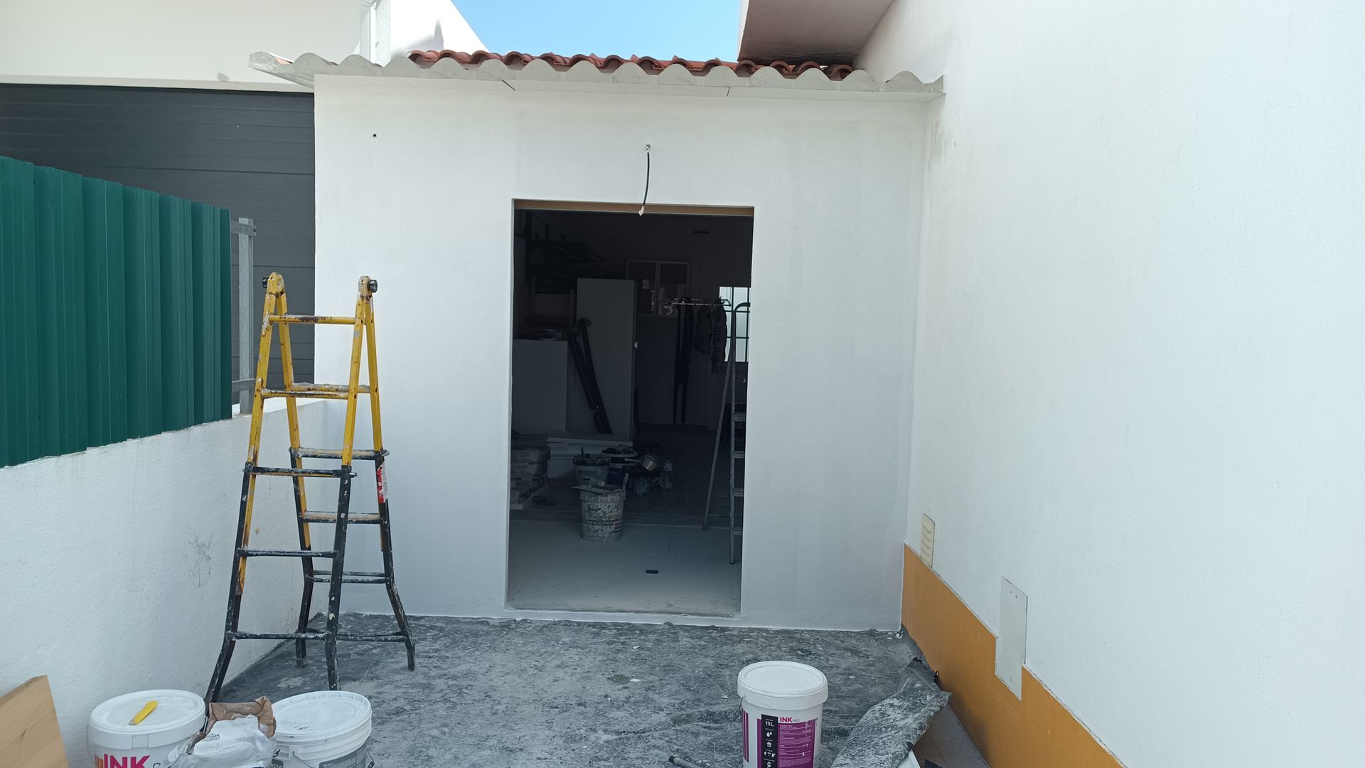 Porta externa em construção, paredes brancas, escada amarela. Interior visível, baldes, ferramentas.