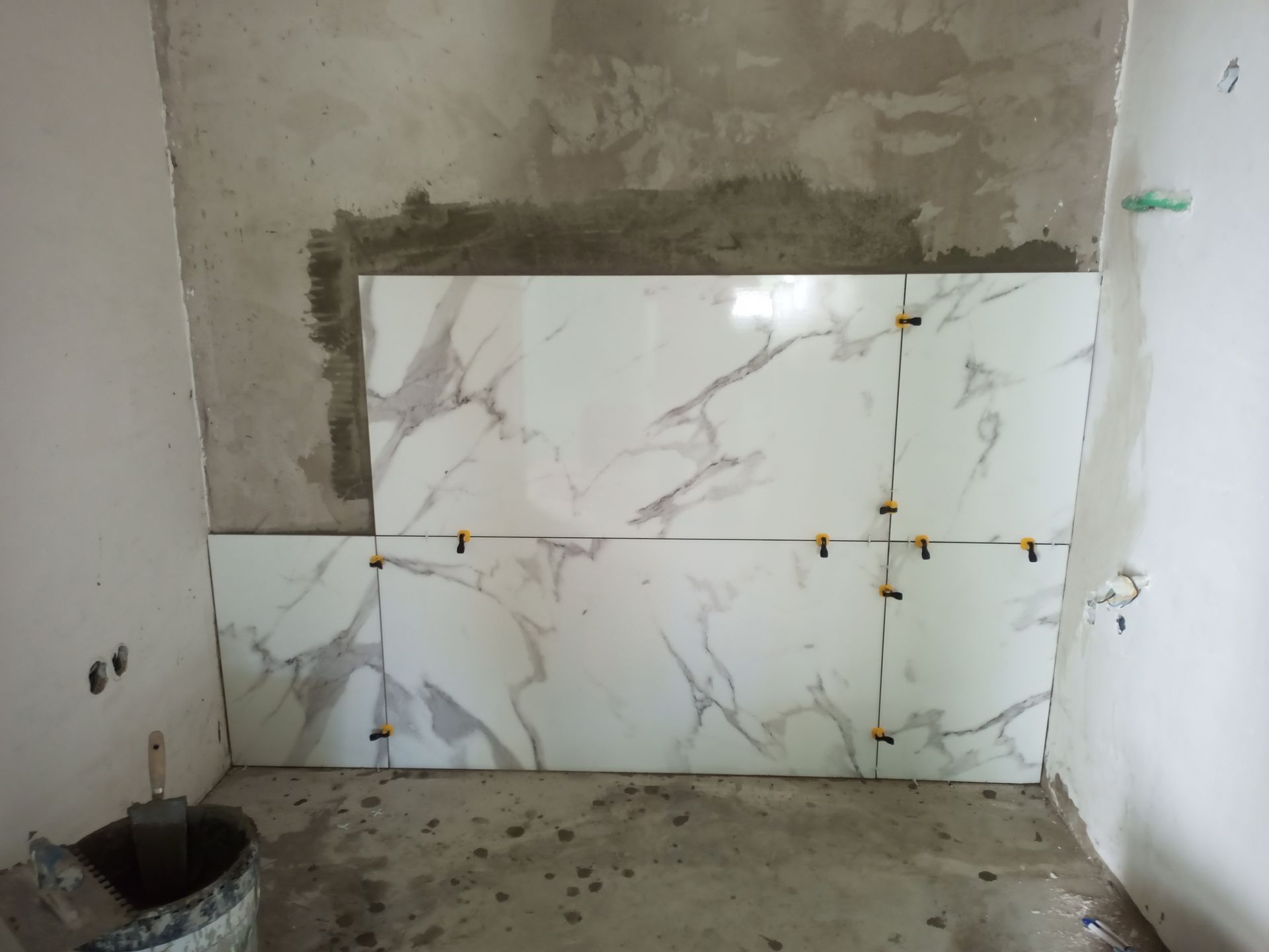 Parede do banheiro em construção, com colocação de azulejos em andamento. Azulejos brancos com aparência de mármore e espaçadores para nivelamento. Paredes de concreto.