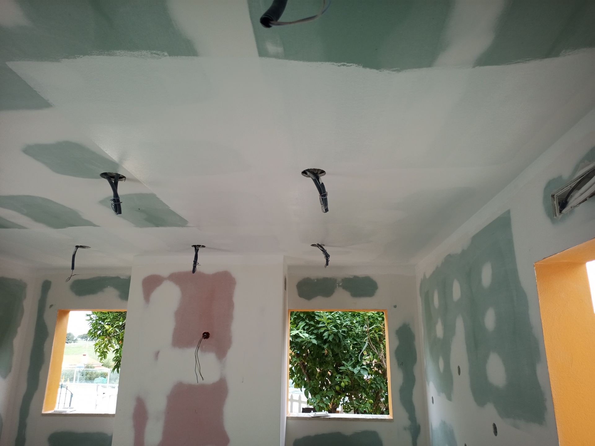 Vista interna de um cômodo em construção com paredes de gesso e fiação exposta; duas janelas mostram vegetação.