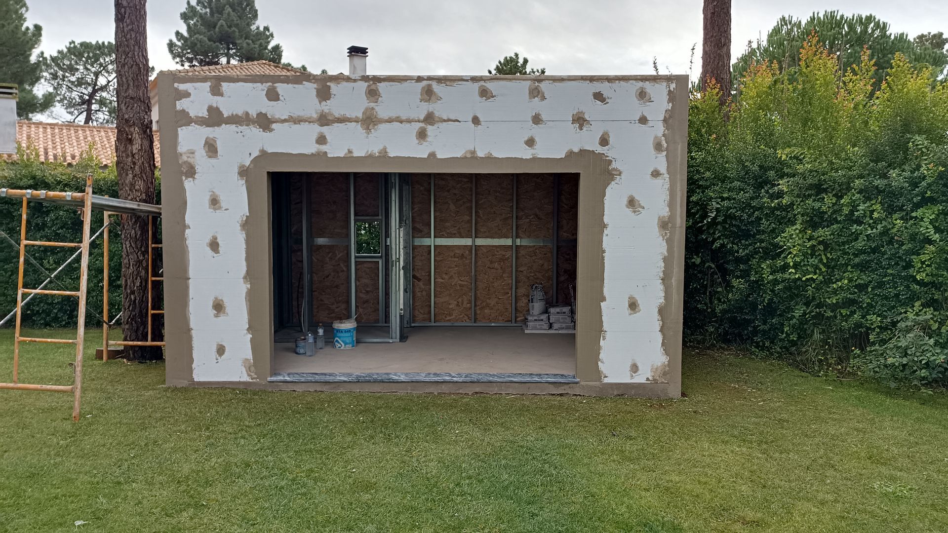 Uma pequena construção inacabada com uma grande abertura, em um gramado. Há um andaime à esquerda.