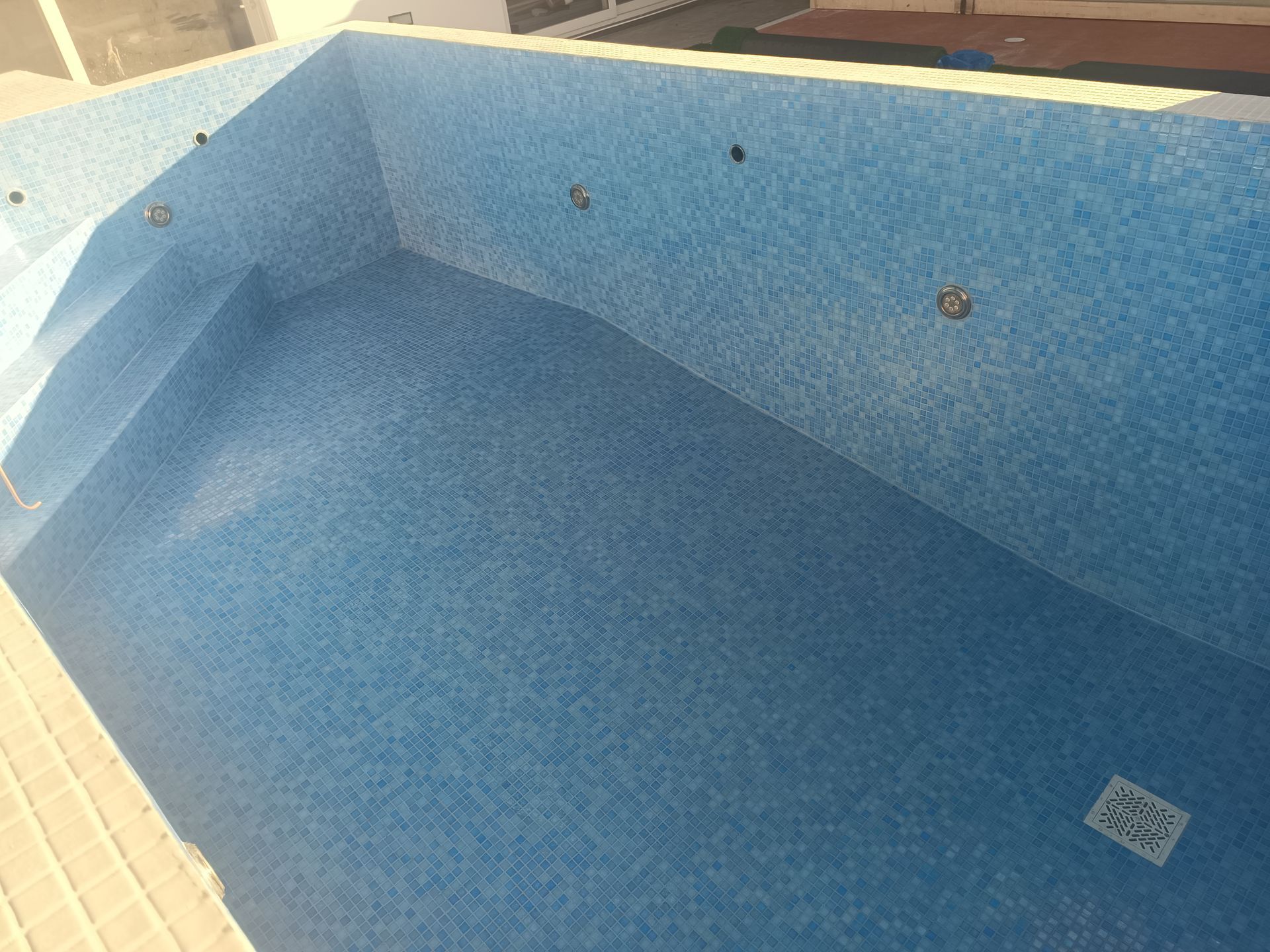 Piscina vazia revestida com azulejos azuis, com degraus e luminárias embutidas, vista de um ângulo superior.