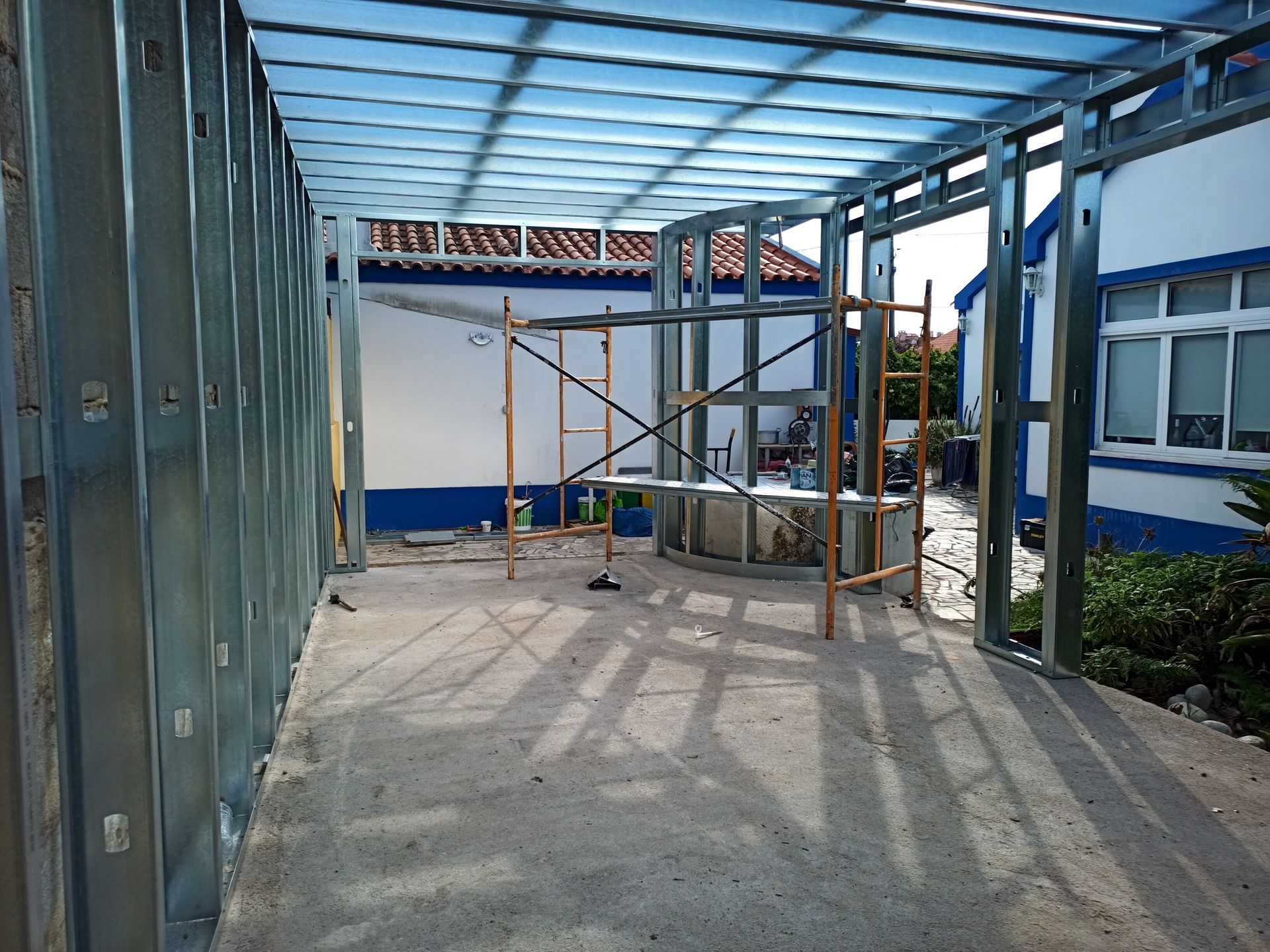 Estrutura metálica de uma sala em construção, piso de concreto, andaimes presentes, vista de um edifício ao fundo.