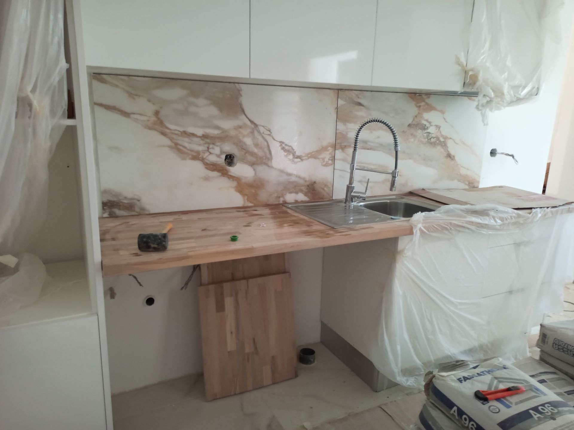 Cozinha em construção: backsplash de mármore, bancada de madeira, pia de aço inoxidável. Armários brancos e revestimento plástico.