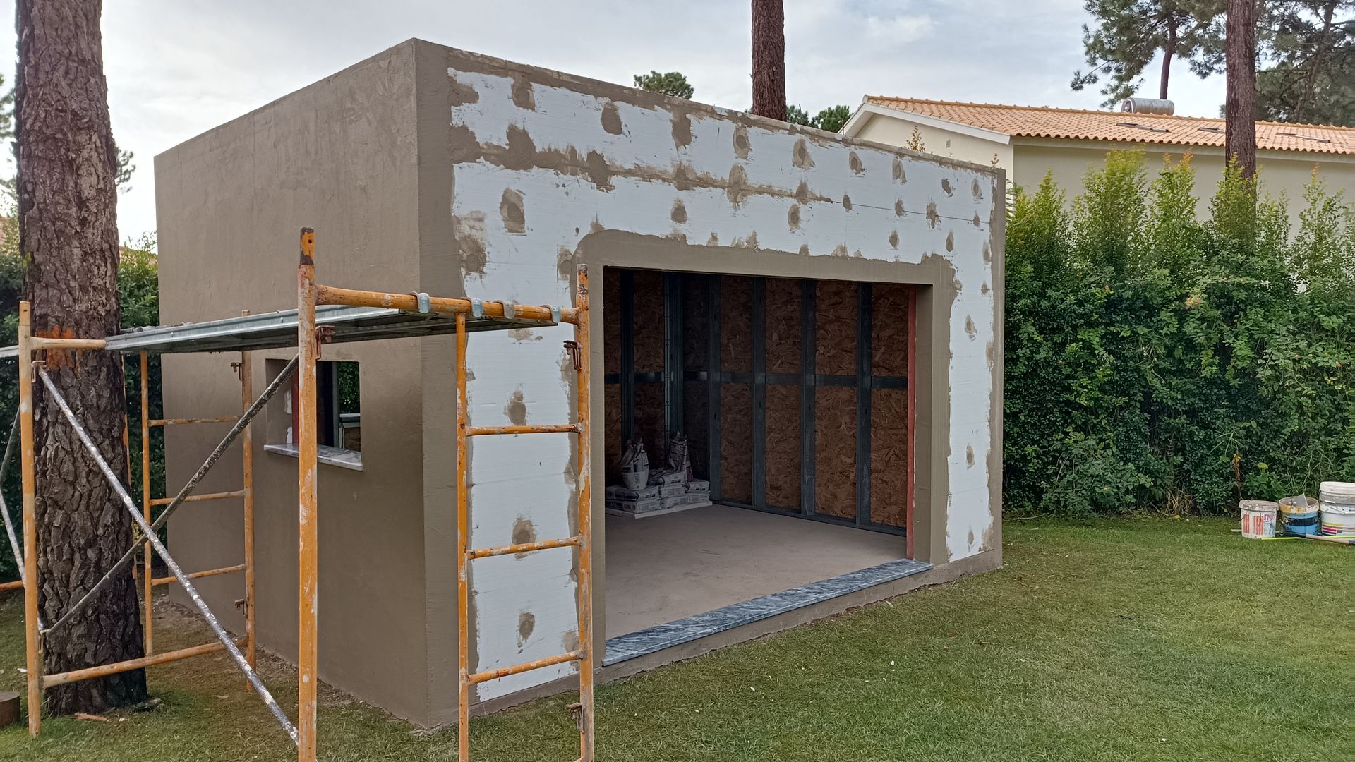 Um pequeno prédio em construção, em tons de bege e branco, com uma porta de garagem aberta, cercado por grama e árvores.