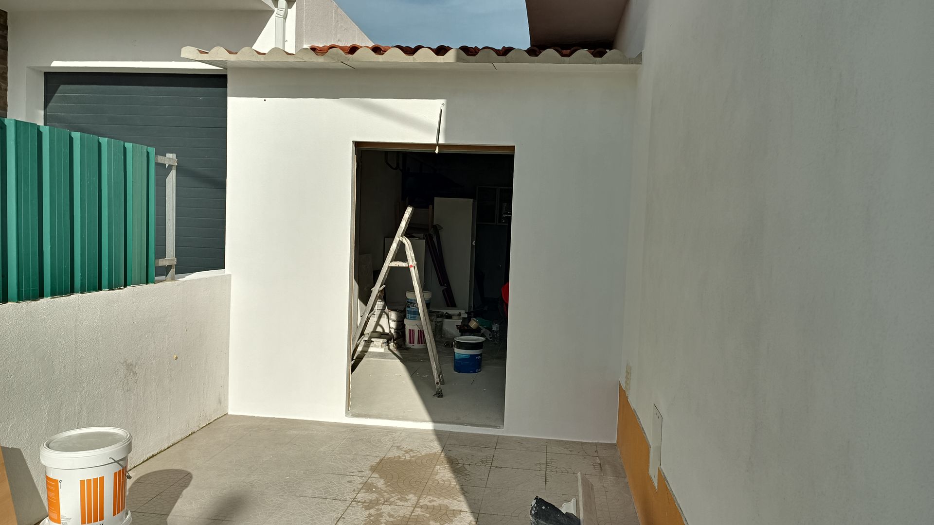 Porta do galpão pintada de branco, escada dentro, cerca verde à esquerda, paredes externas também brancas, chão de concreto.