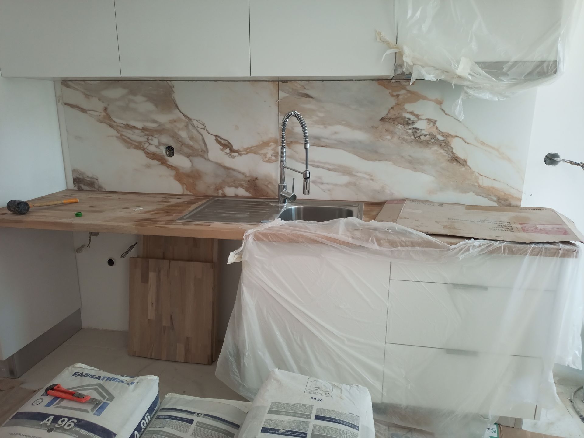 Cozinha em construção com backsplash de mármore, pia de aço inoxidável, armários brancos e bancada de madeira.