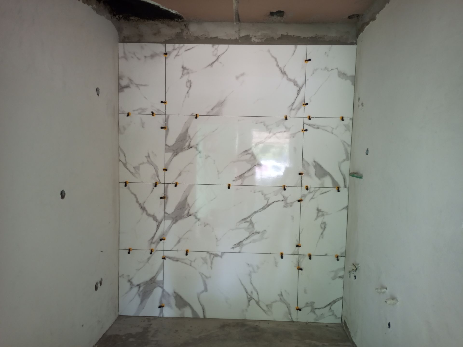Azulejos brancos com aparência de mármore instalados em uma parede de um cômodo em construção, com espaçadores visíveis.