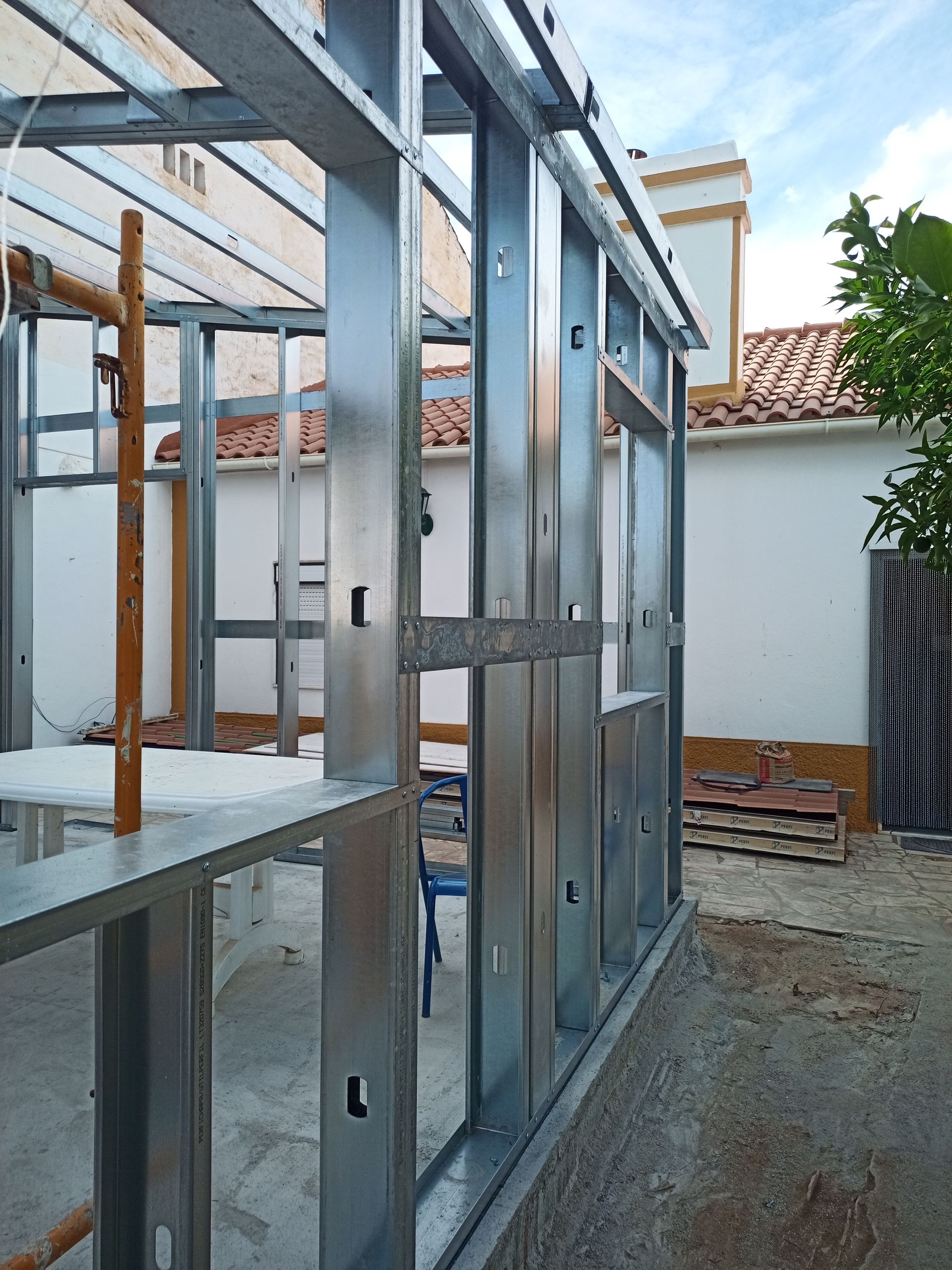 Estrutura metálica para a parede externa de um edifício em construção. Canteiro de obras com prédio branco e céu azul ao fundo.