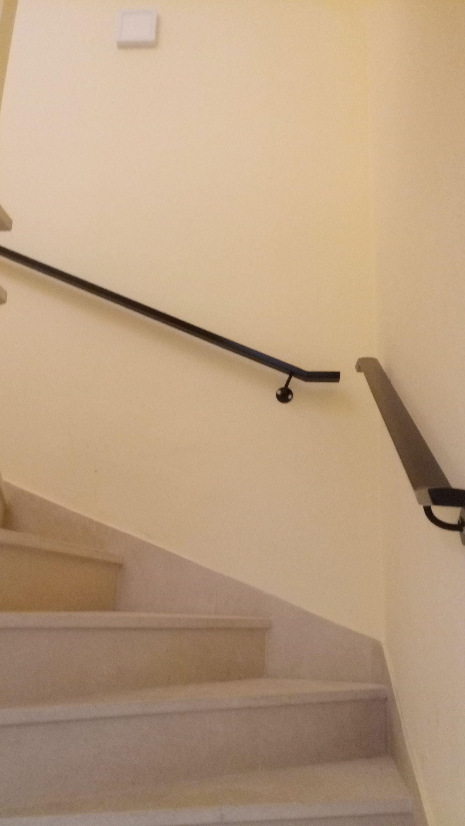 Corrimão de metal preto em uma parede bege ao lado de uma escada com degraus claros.
