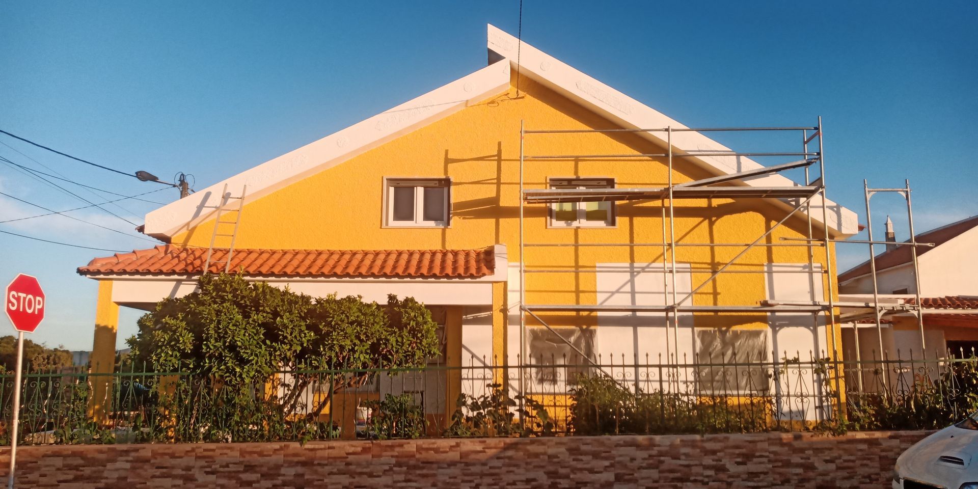 Casa amarela com andaimes, telhado de telha vermelha, placa de pare e céu azul.