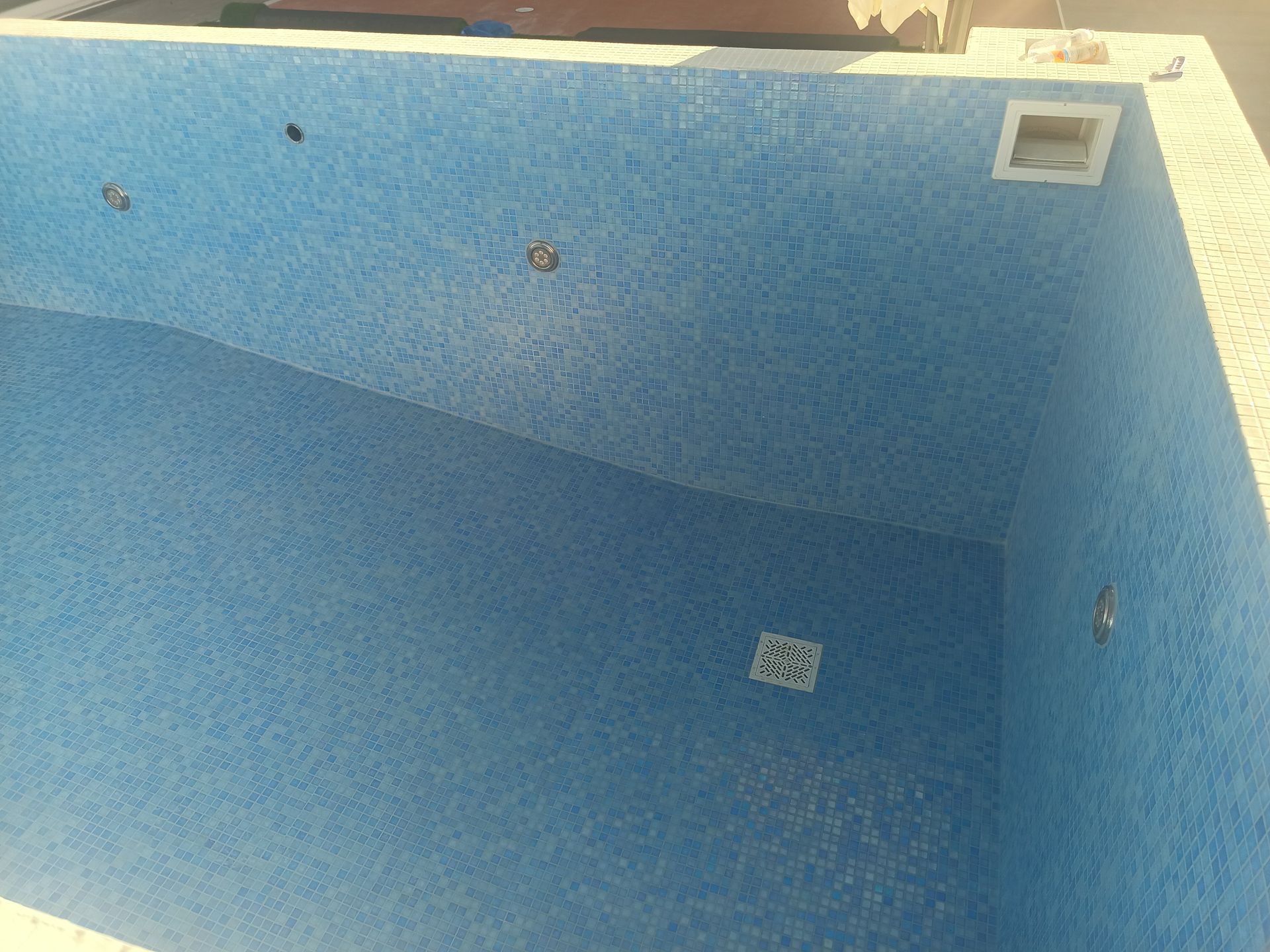 Piscina vazia revestida com azulejos azuis. Ralo quadrado e entrada de água visíveis. Luz solar.