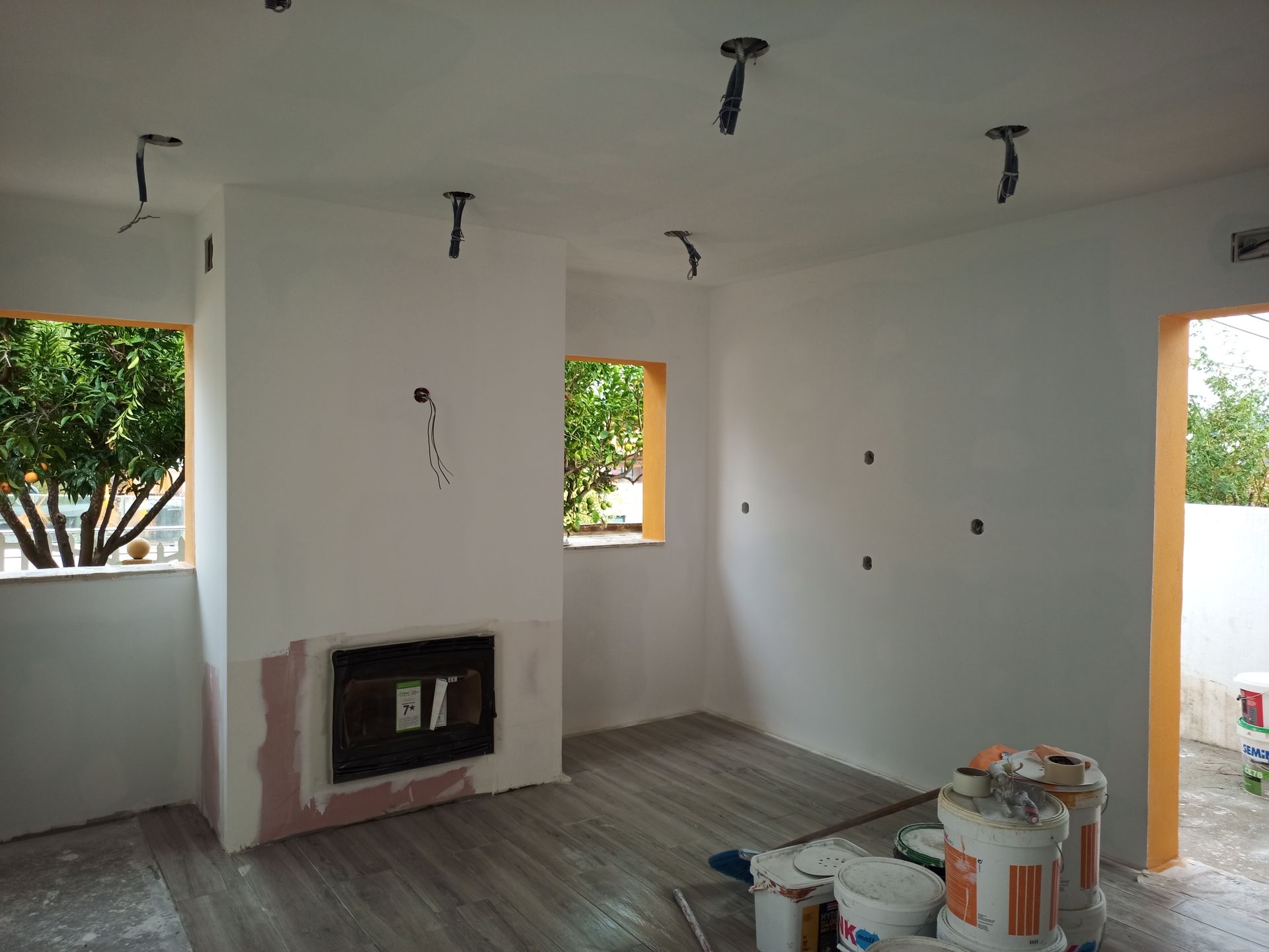 Interior de um cômodo em construção, com paredes brancas, fiação inacabada e janelas com molduras laranja.