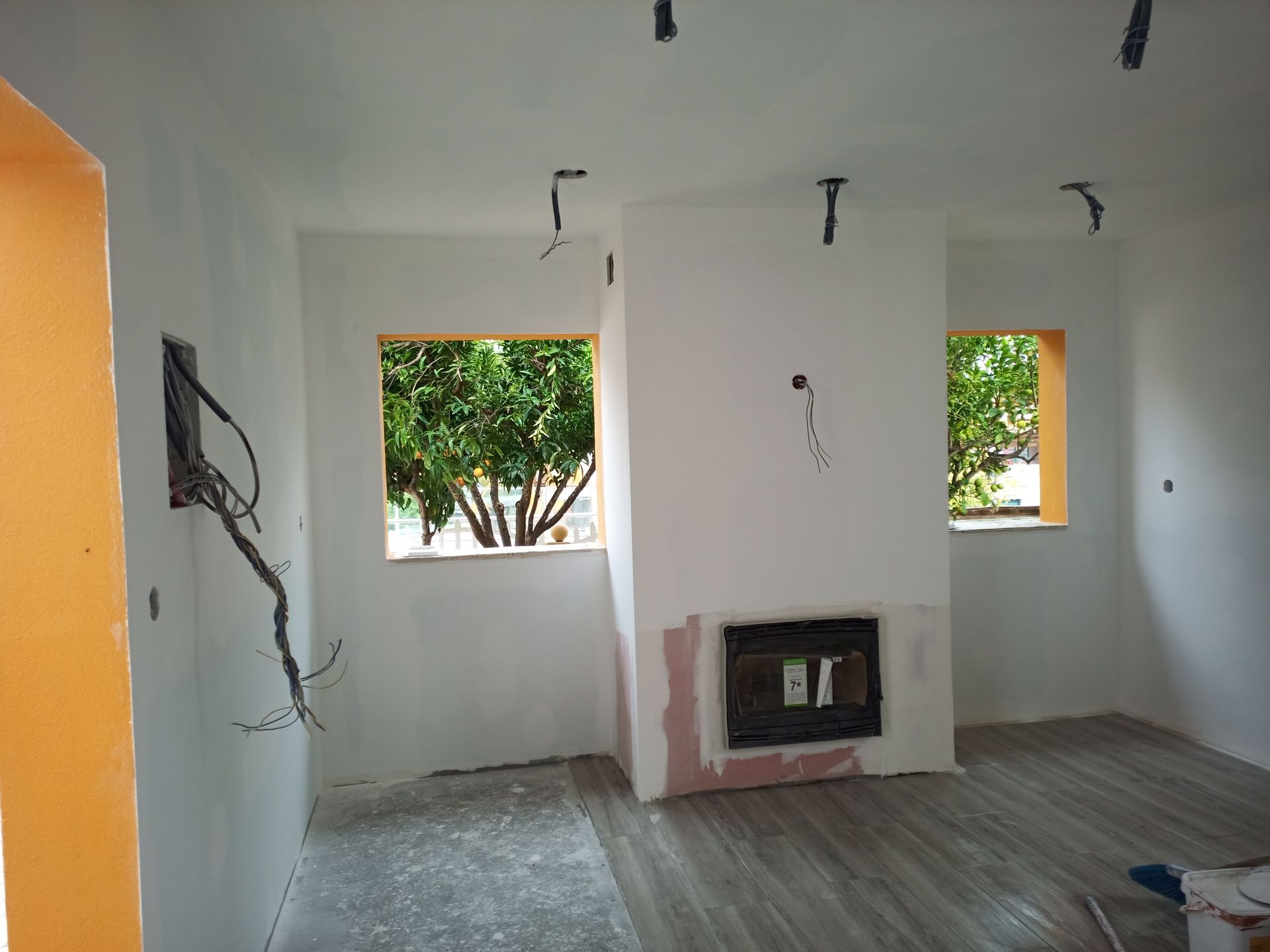 Quarto interior em construção com paredes brancas, janelas, lareira e fiação exposta.