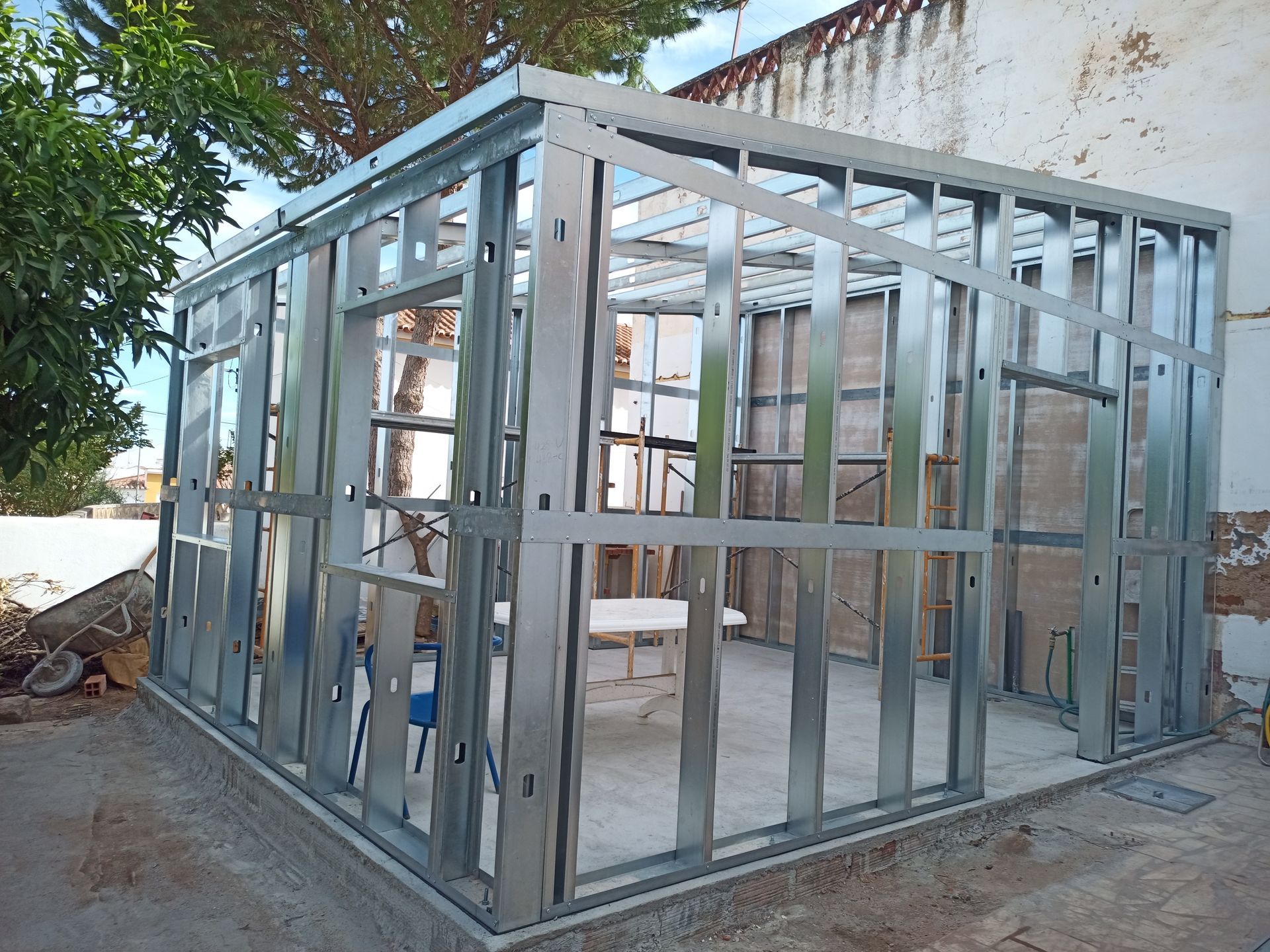 Estrutura metálica de um edifício em construção, ao ar livre, contra uma parede parcialmente danificada e árvores.