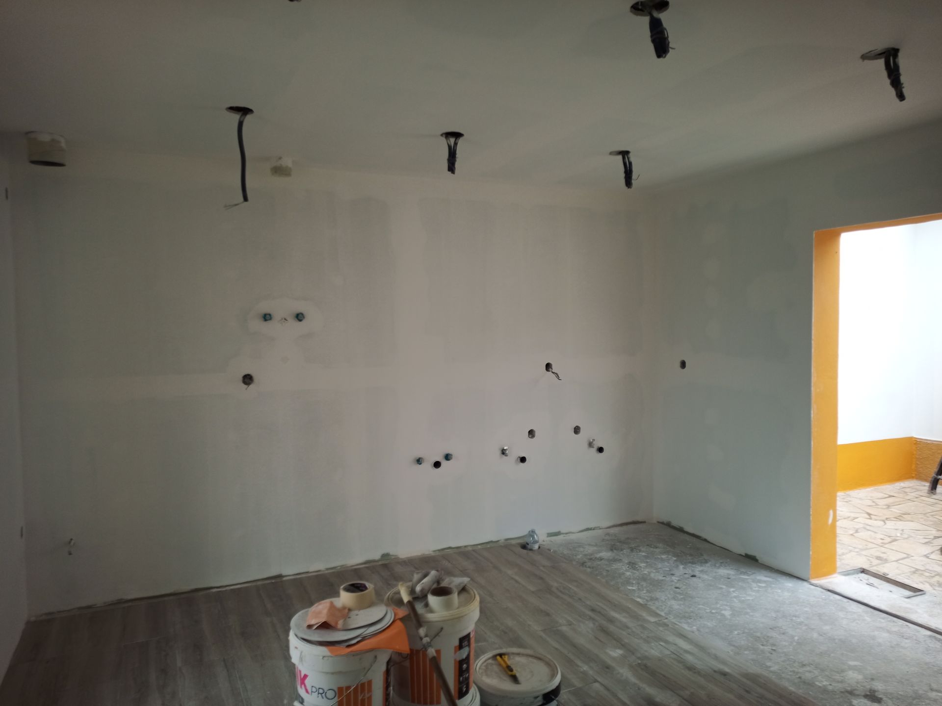 Quarto interior com paredes brancas, fiação exposta, luminárias e baldes de tinta no chão.