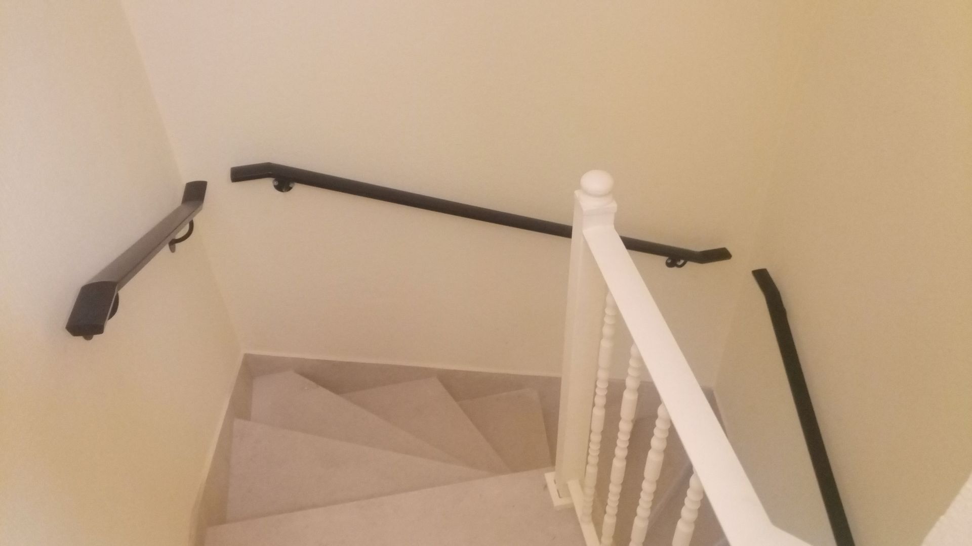 Escadaria com degraus brancos, paredes creme e corrimãos pretos, com um balaústre branco à direita.