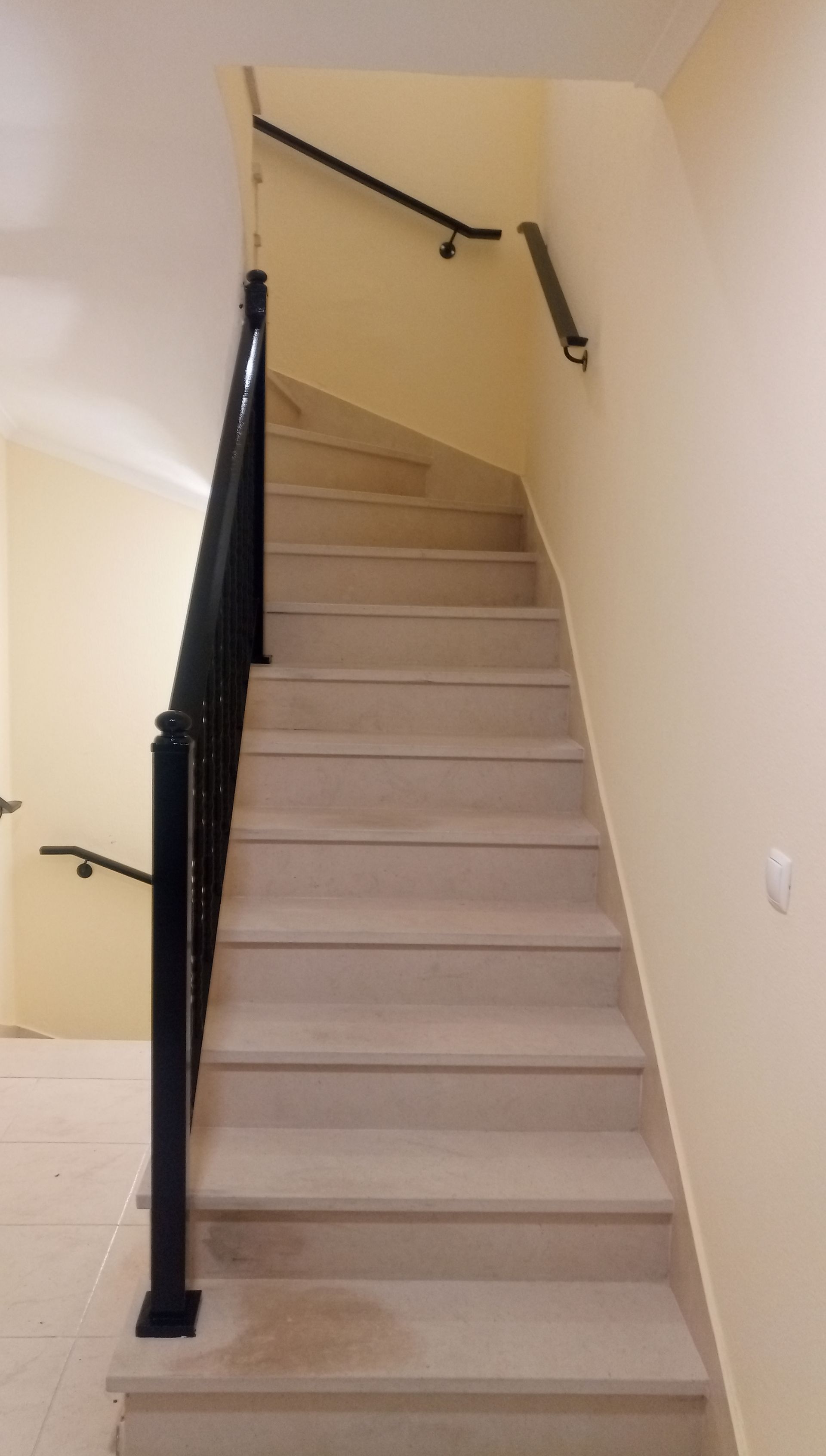 Escada com degraus claros e corrimão preto. As paredes são bege. Um corrimão percorre a parede lateral.