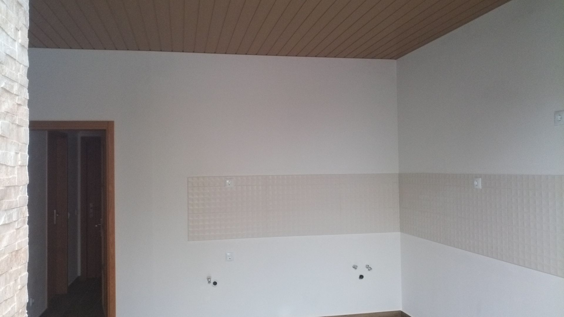 Vista interior de um cômodo com paredes brancas, teto bege e porta de entrada, com revestimento externo em azulejo e pedra.