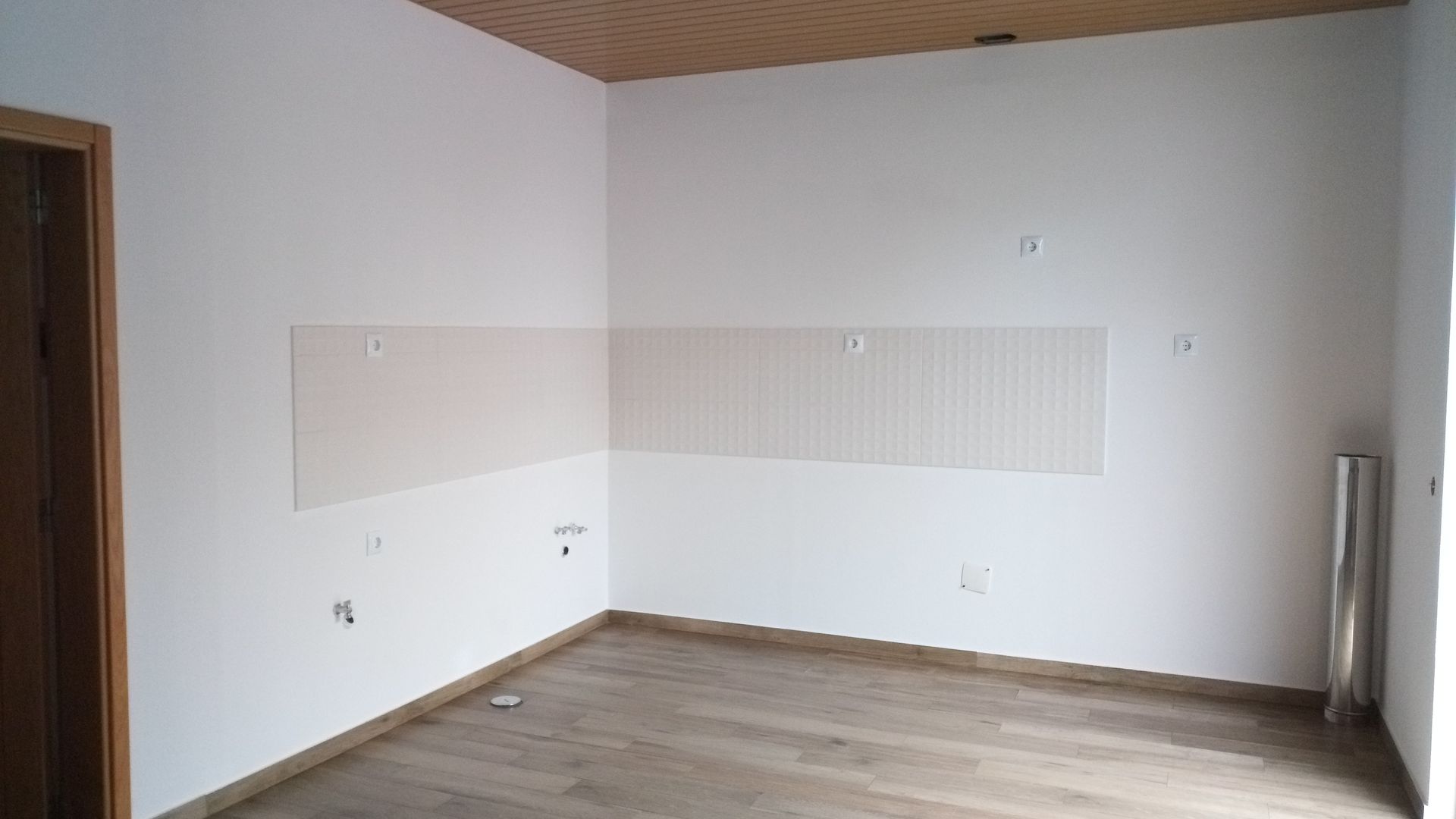 Quarto vazio com piso e teto de madeira, paredes brancas e porta à esquerda.