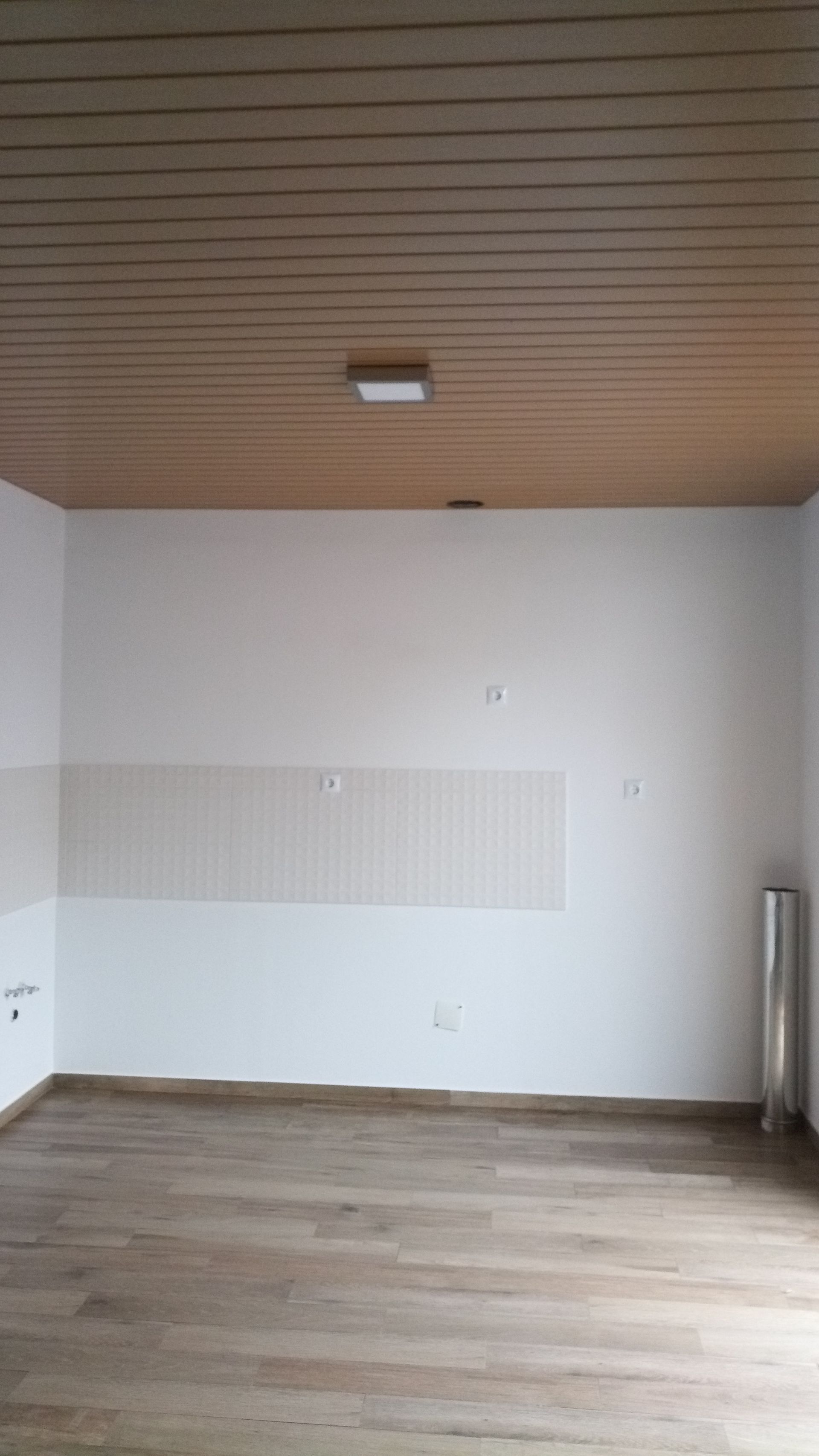 Sala vazia com teto revestido de madeira, paredes brancas e piso que imita madeira. Uma única lâmpada está acesa.