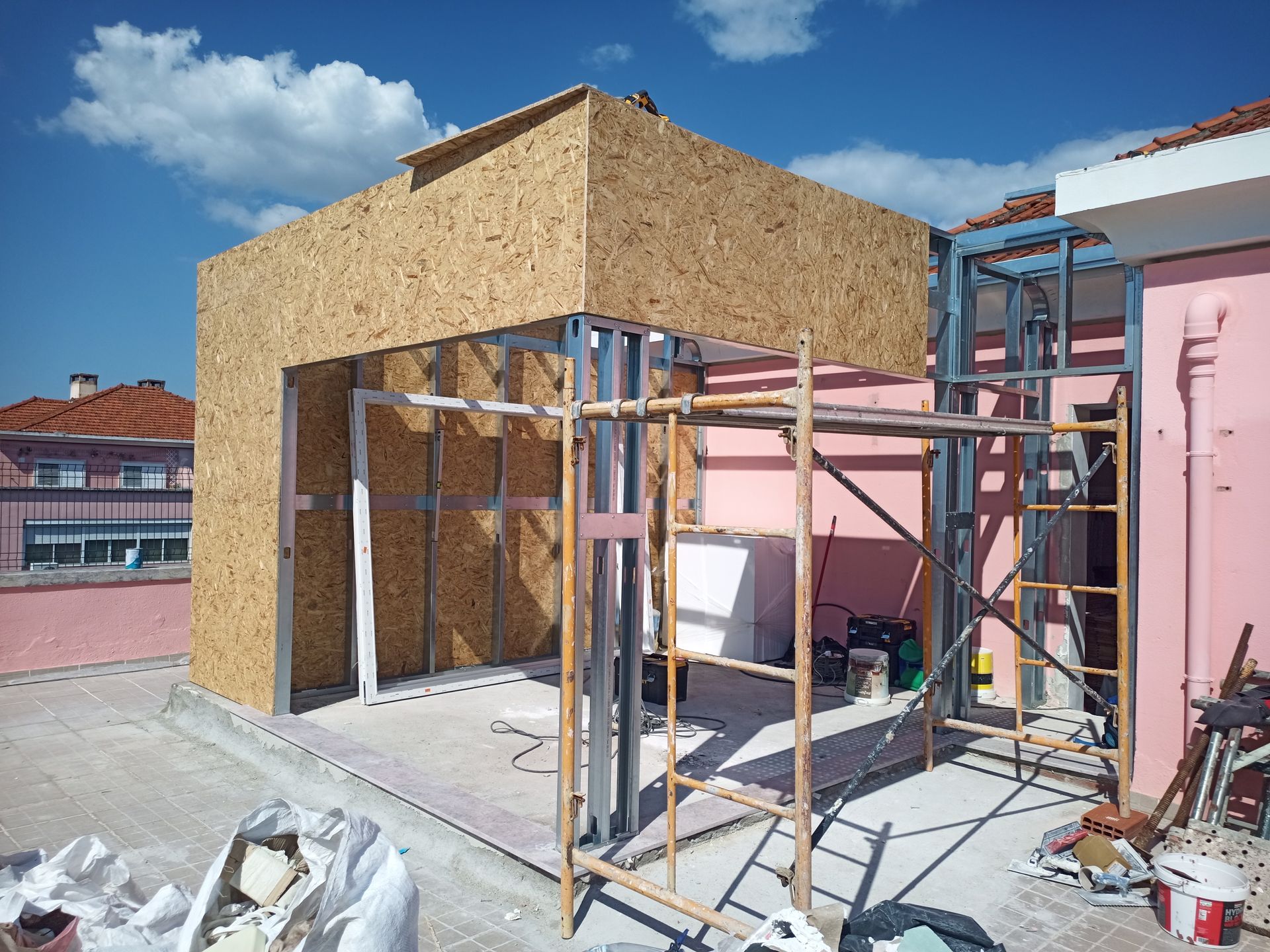 Construção de um anexo em um telhado rosa, estrutura metálica parcialmente revestida com painéis de madeira, dia ensolarado.