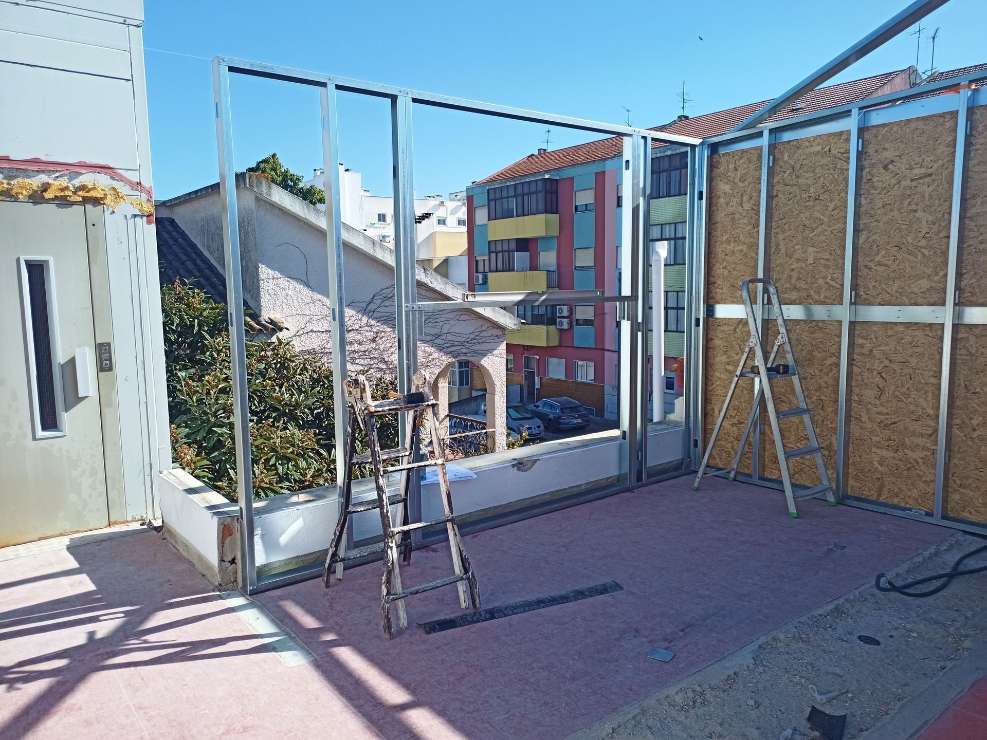 Canteiro de obras com estrutura metálica, painéis de madeira e escadas em uma varanda com vista para edifícios urbanos.