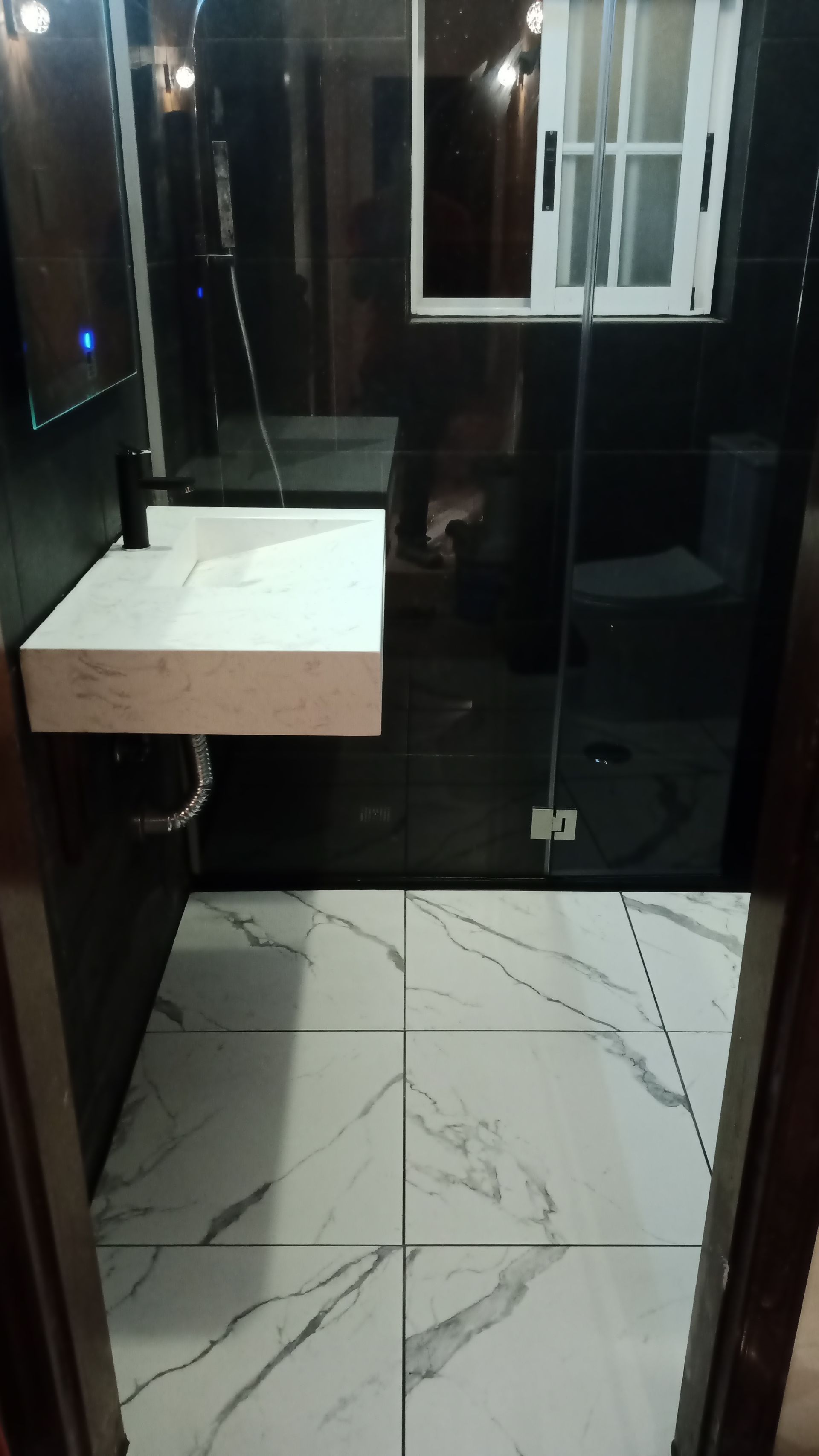 Banheiro moderno com paredes revestidas de azulejos pretos, piso com aparência de mármore branco, pia suspensa branca e box de vidro.