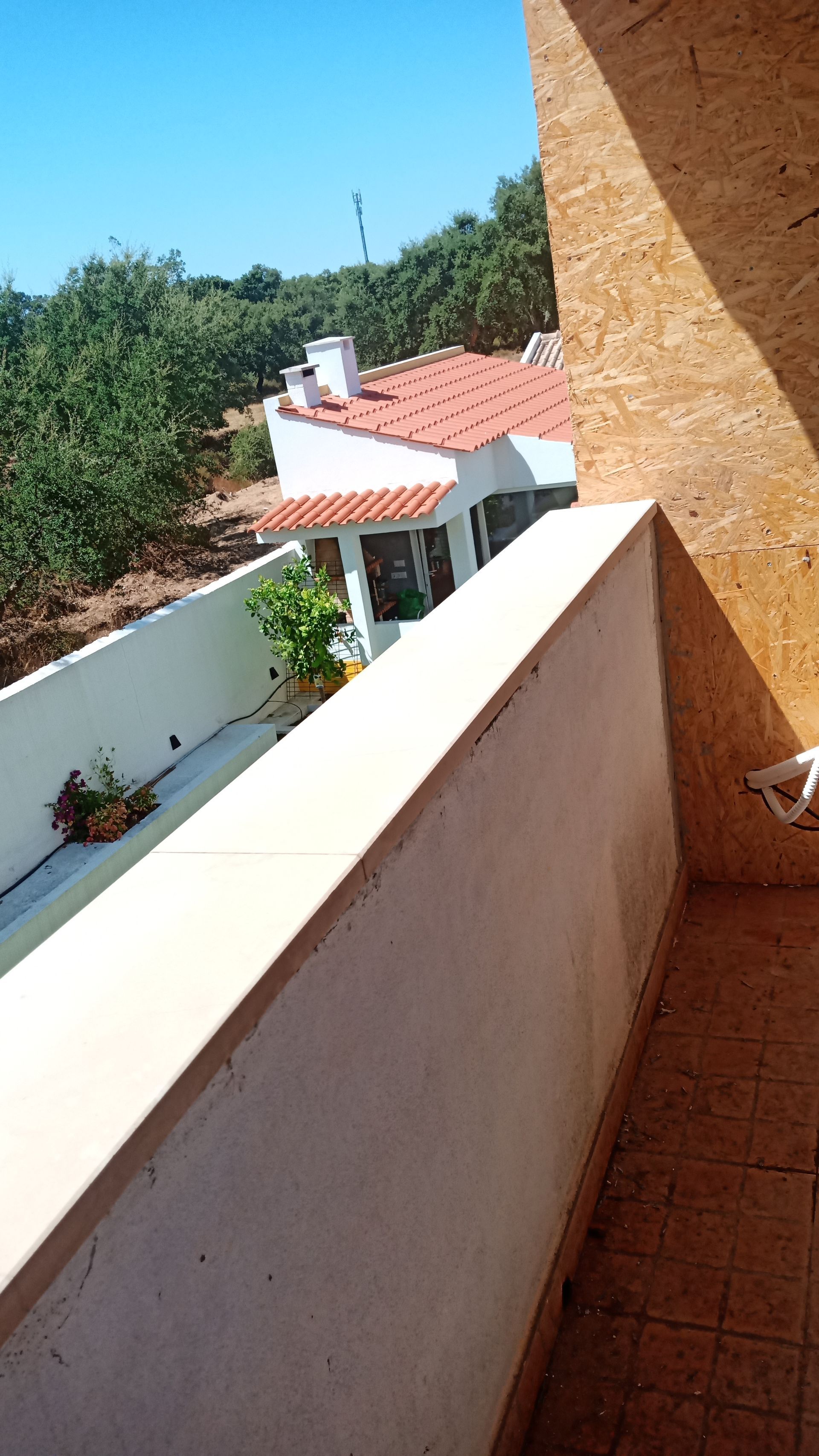 Vista de uma varanda com vista para um prédio branco com telhado laranja e árvores em um dia ensolarado.