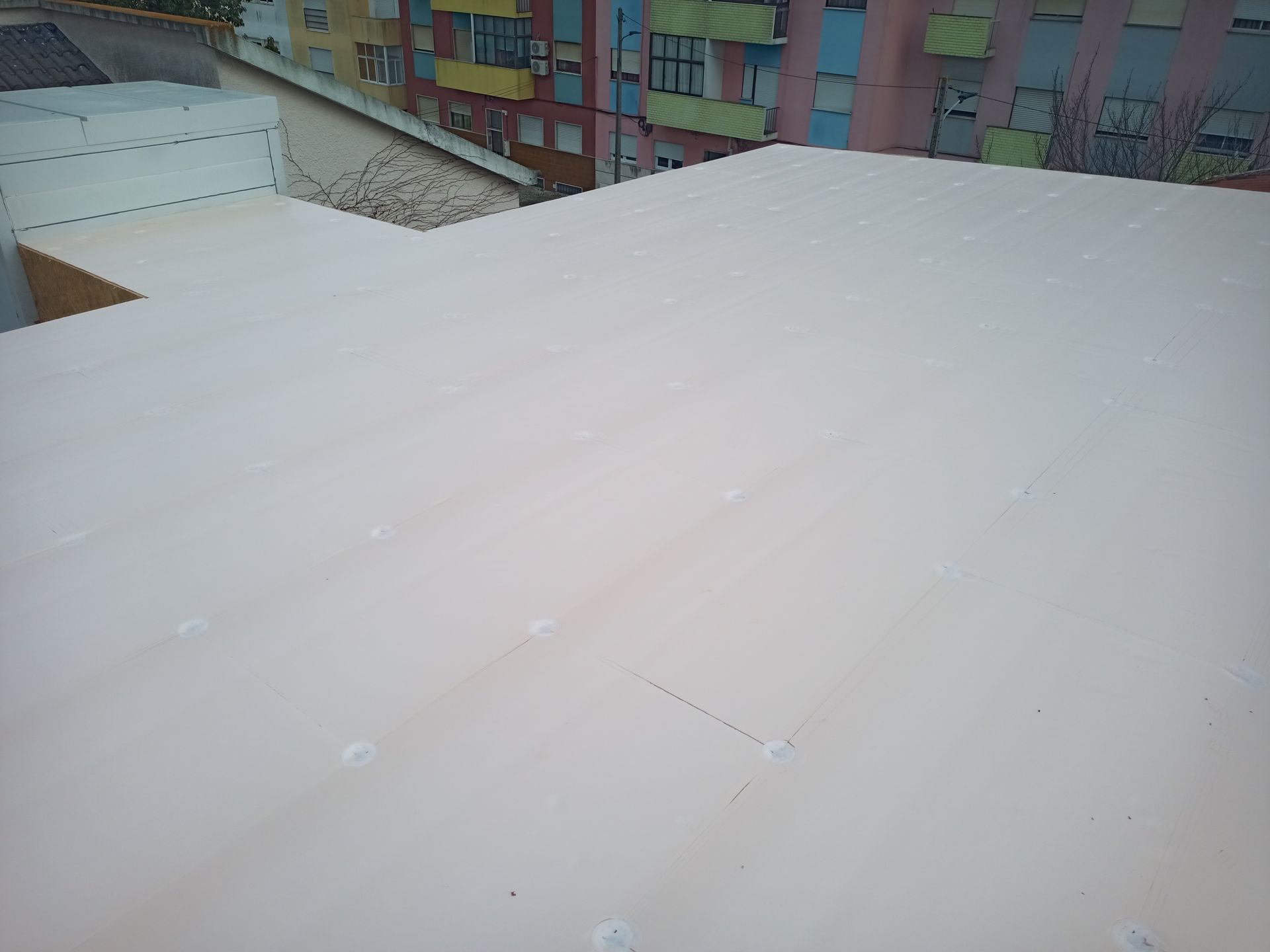 Telhado de metal ondulado rosa claro em um prédio, visto de cima. Prédios coloridos ao fundo.