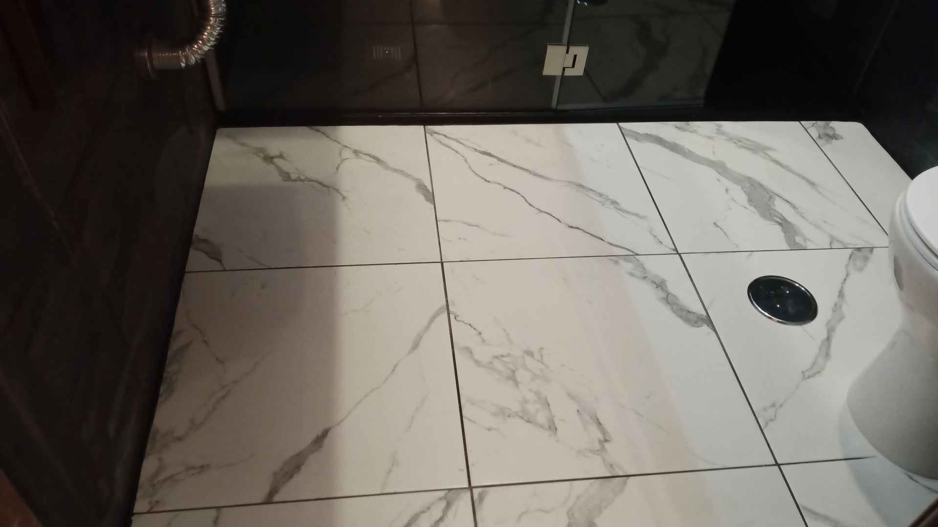 Piso do banheiro com azulejos brancos com estampa de mármore e rejunte preto, com o vaso sanitário visível à direita.