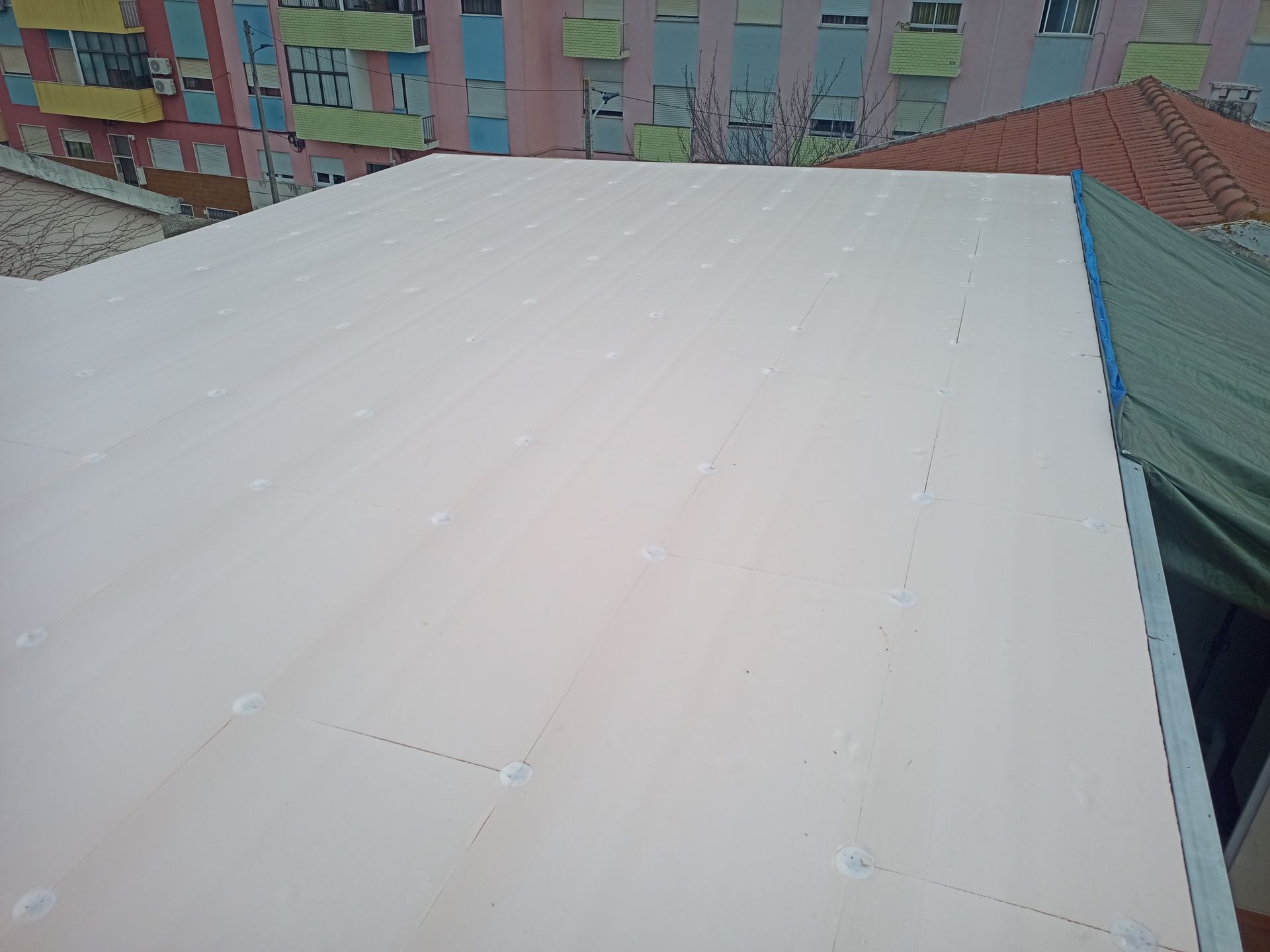 Telhado de metal ondulado branco com fixadores visíveis, vista parcial dos edifícios circundantes.