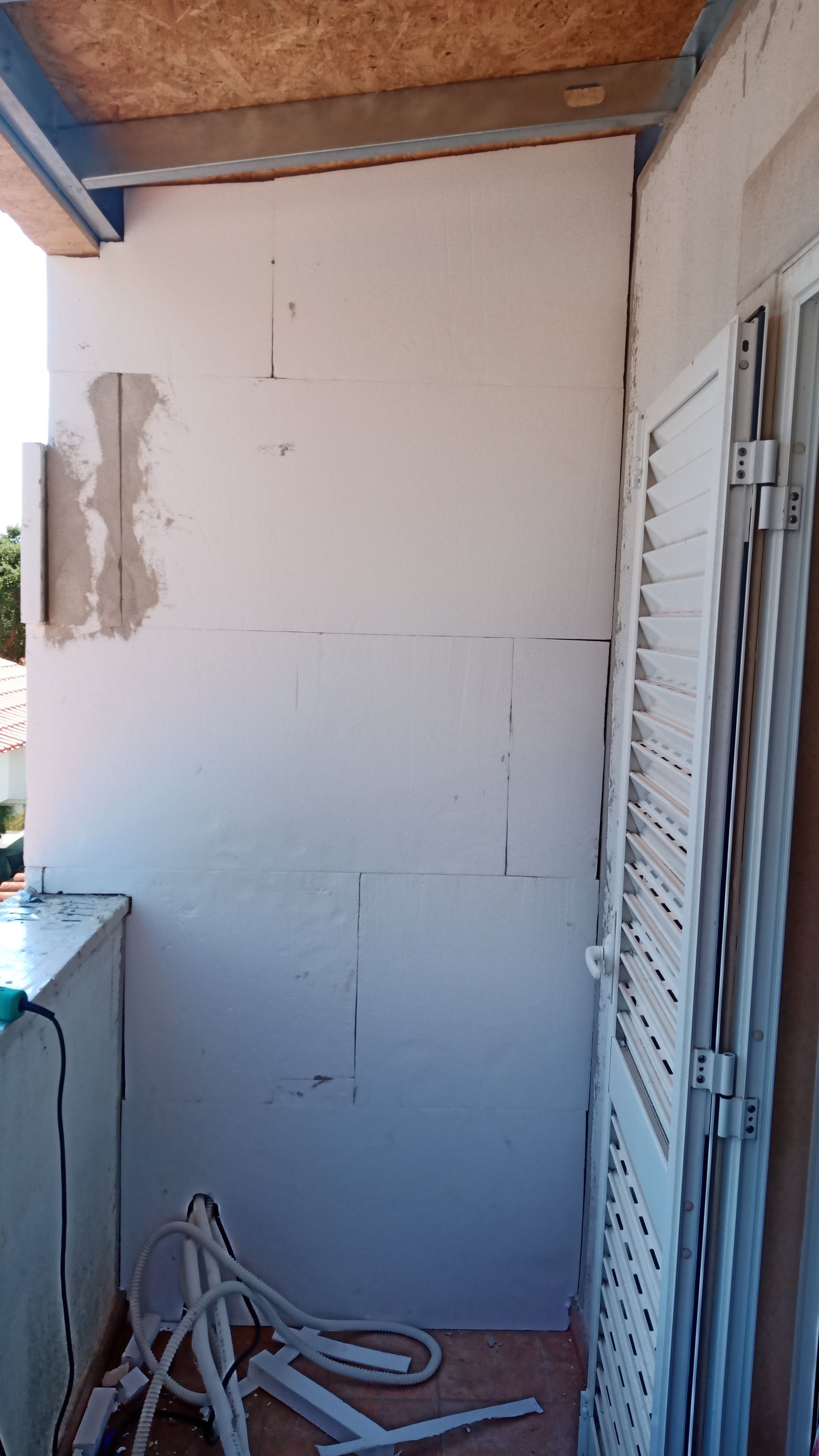 Construção da parede da varanda com isolamento de espuma branca, reboco inacabado e persianas brancas fechadas.