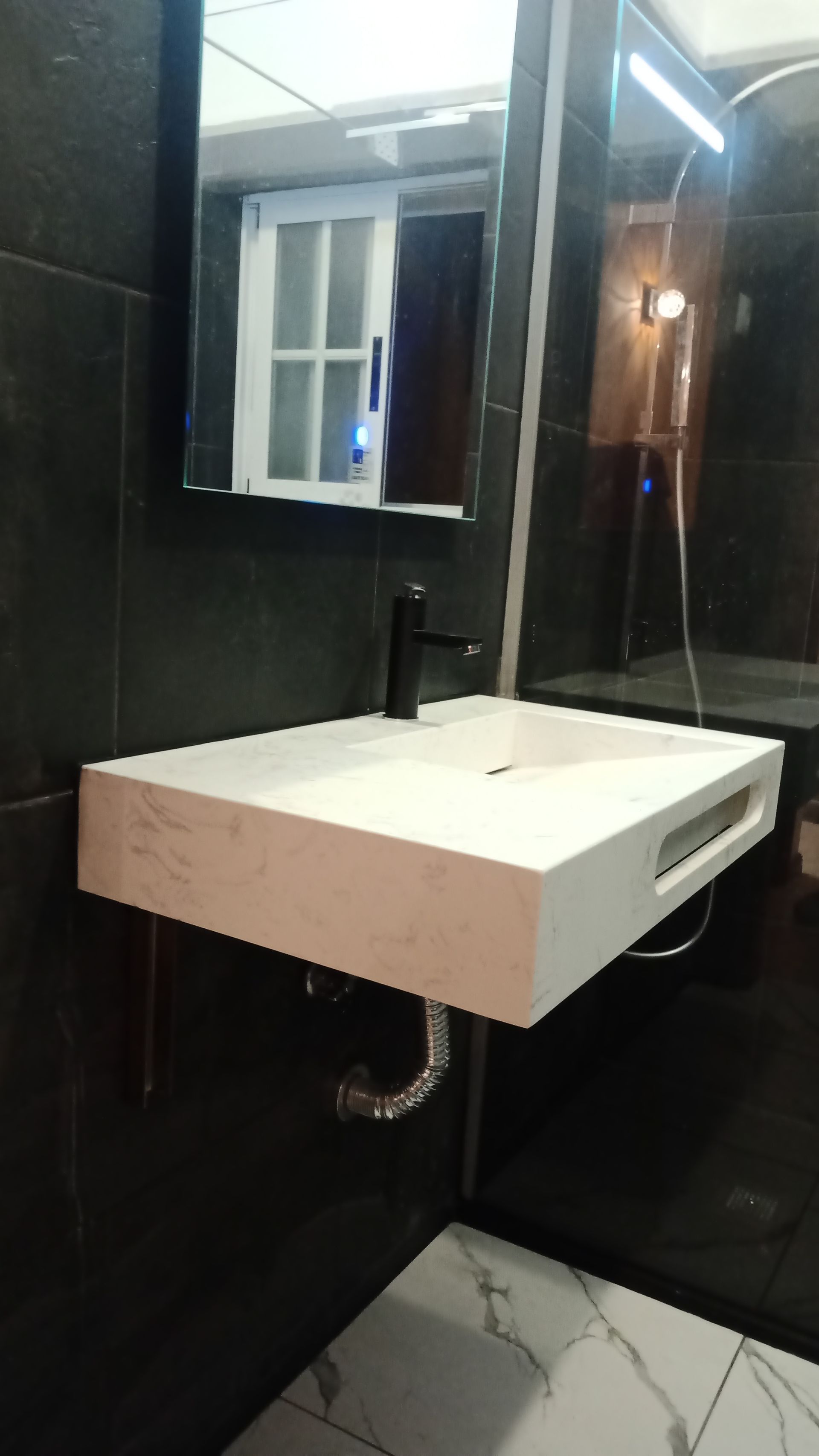 Pia suspensa de mármore branco com torneira preta em parede revestida com azulejos pretos, armário espelhado.