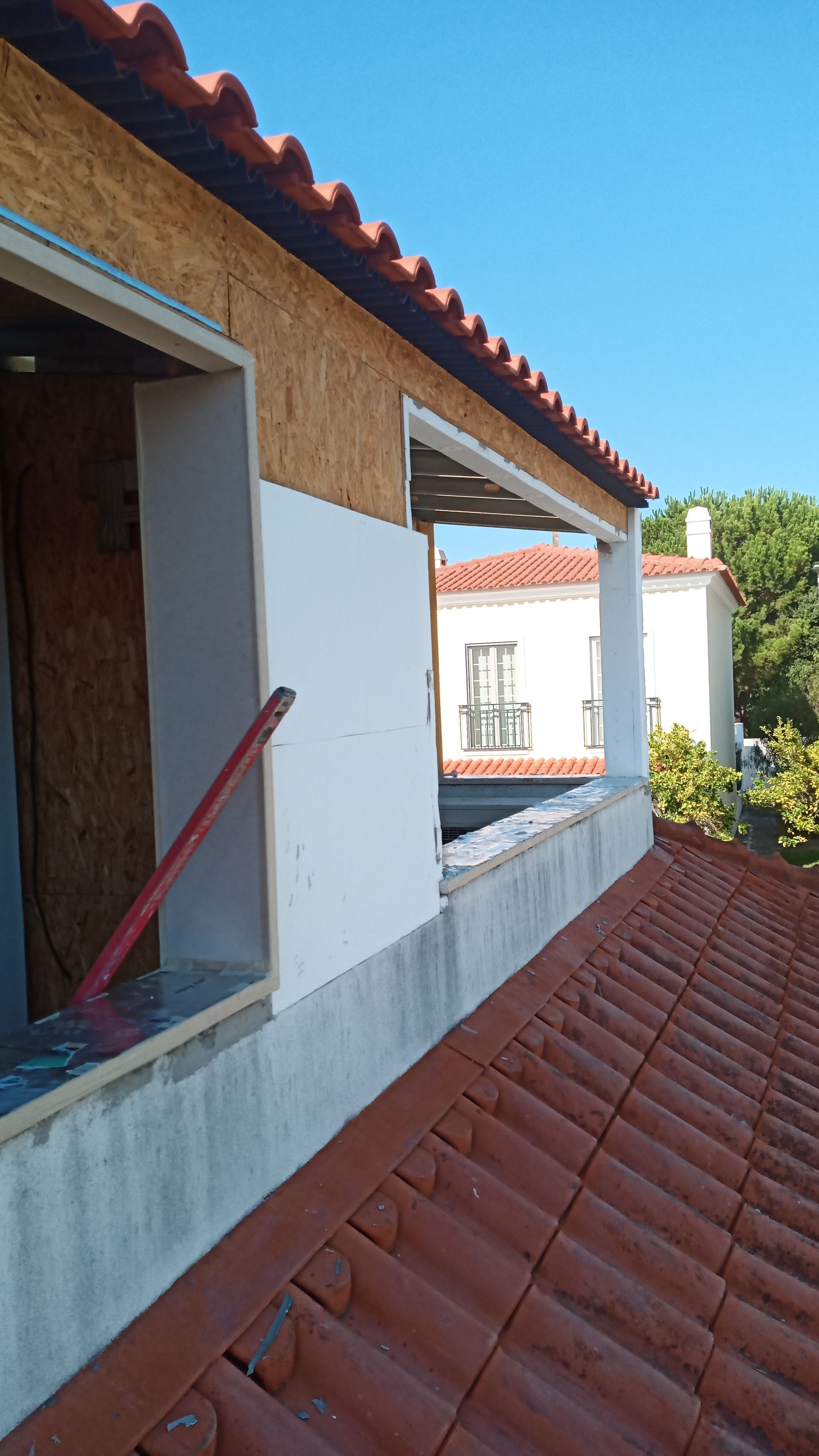 Vista exterior de um edifício em construção, mostrando telhas, uma parede parcialmente construída e um céu azul.