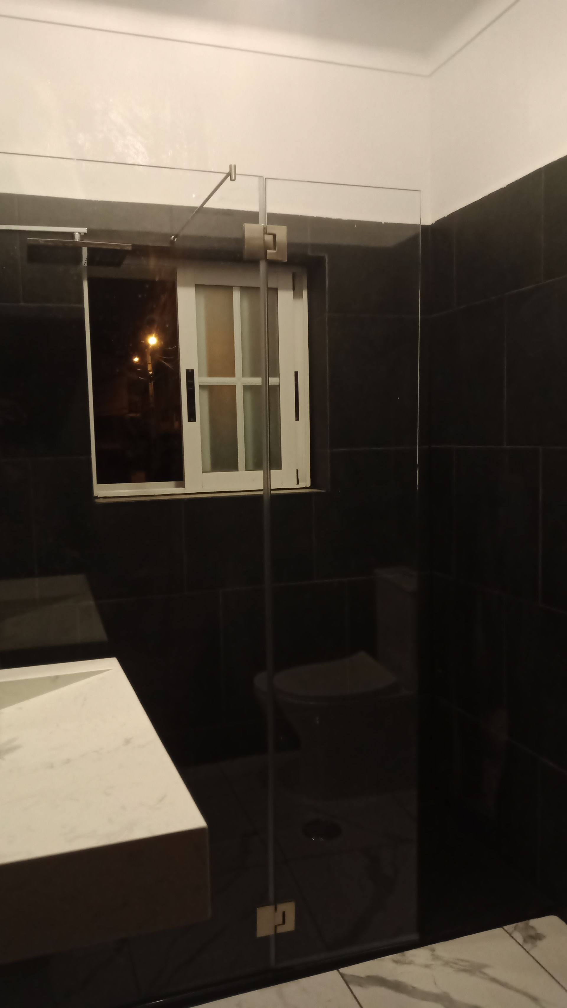 Banheiro com paredes revestidas de azulejos pretos, box de vidro e janela com moldura branca.