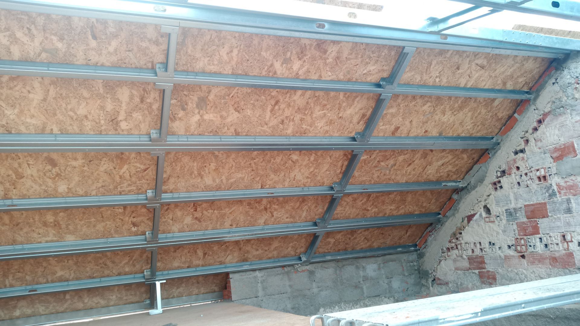Estrutura metálica com painéis de madeira instalada em um teto inclinado; parede de tijolos à direita.